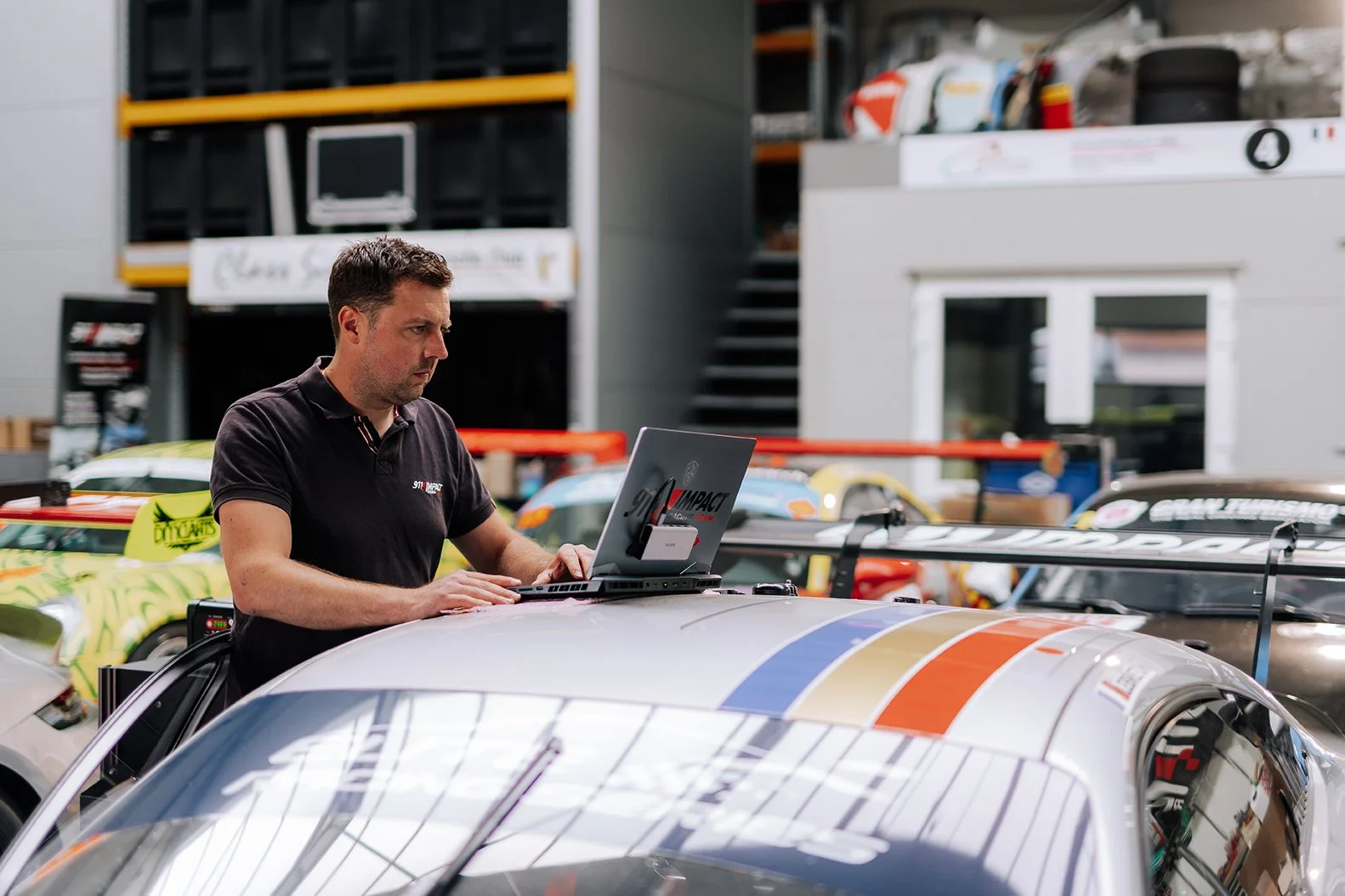 A man working on a racing car with a laptop in a garage or workshop with multiple race cars and equipment in the background.