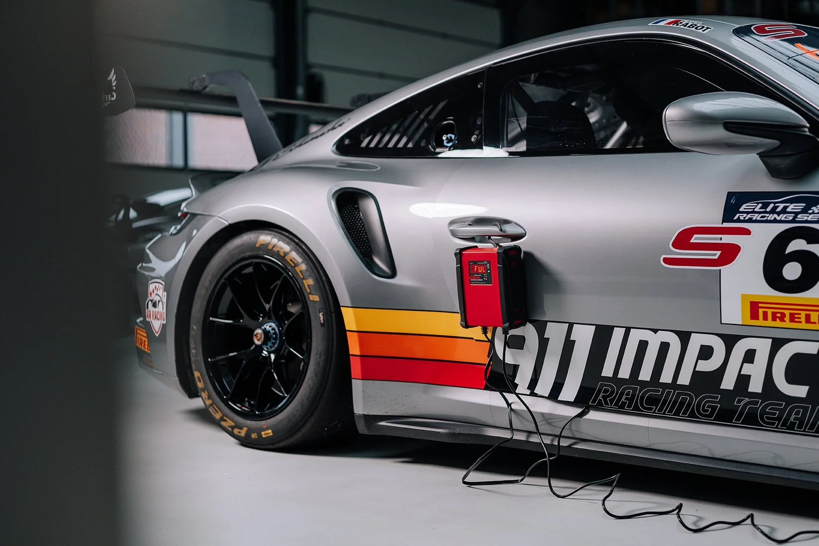 A silver race car with Pirelli tires in a garage, connected to a red electronic device with a digital display. The car has racing decals, including an 'Impact Racing' logo, and features an aerodynamic design with a rear wing.