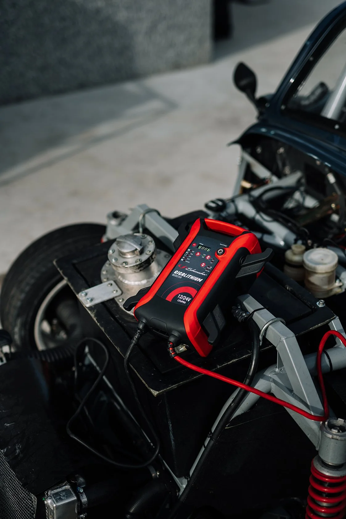 A diagnostic automotive scanner is placed on a car engine, connected with red and black cables, inside a garage or workshop.