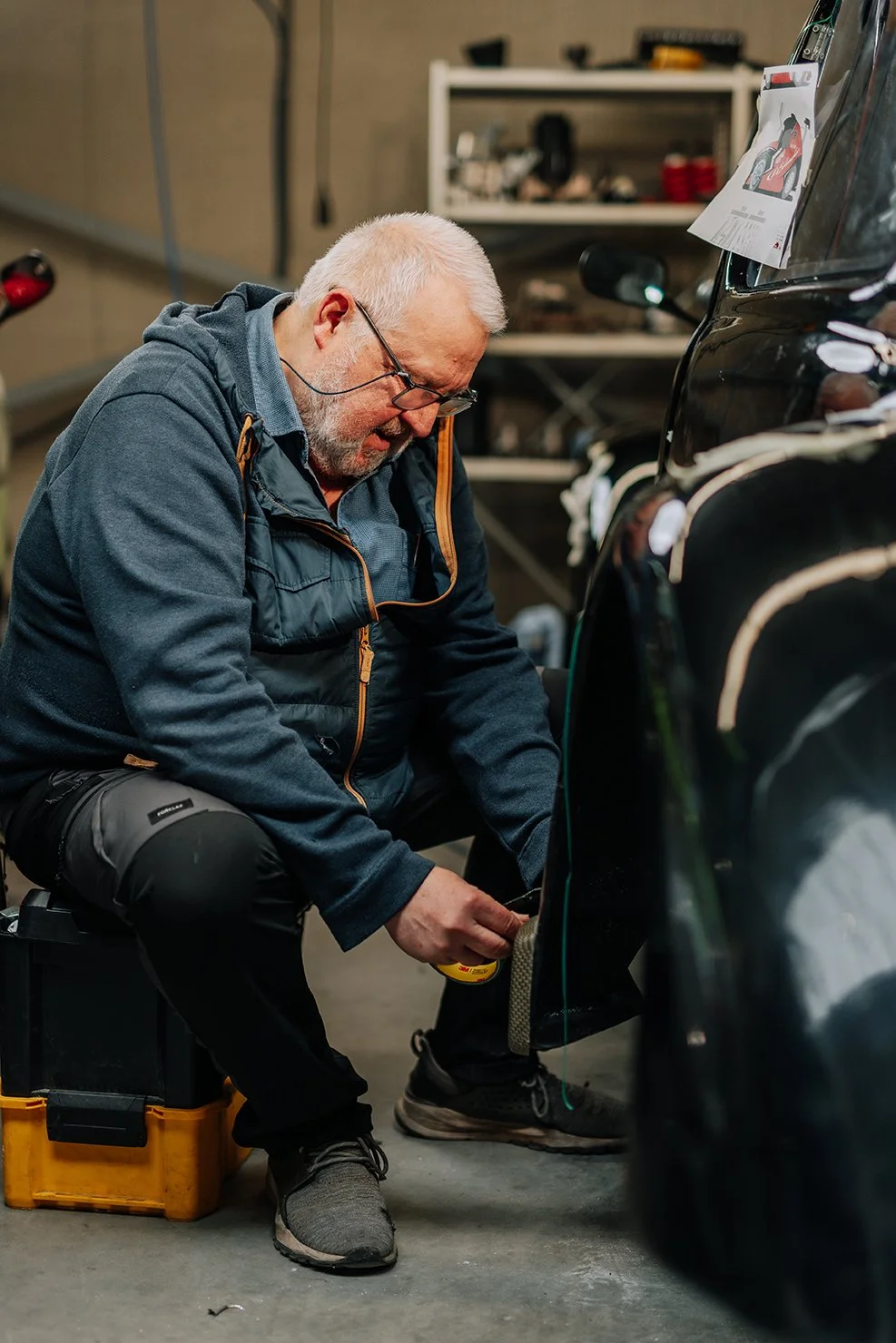 An older man with glasses working on a black car in a garage, sitting on a black and yellow toolbox, focusing on the car's wheel.