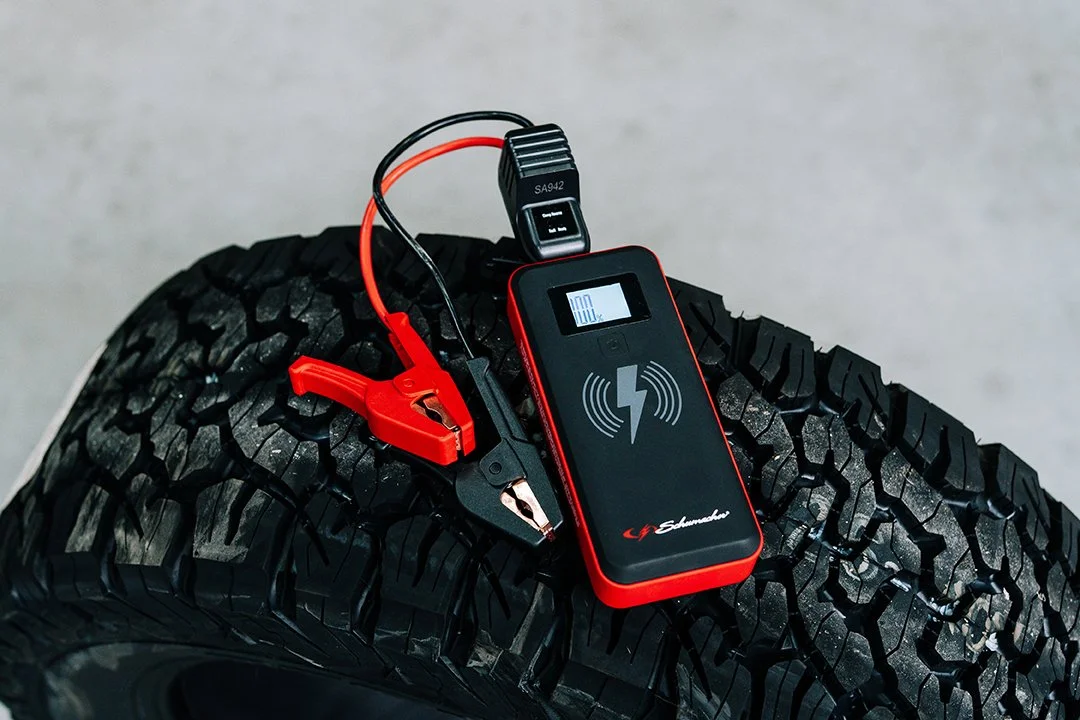A power supply tester connected to a car battery jumper clamp and a digital battery tester resting on a tire.