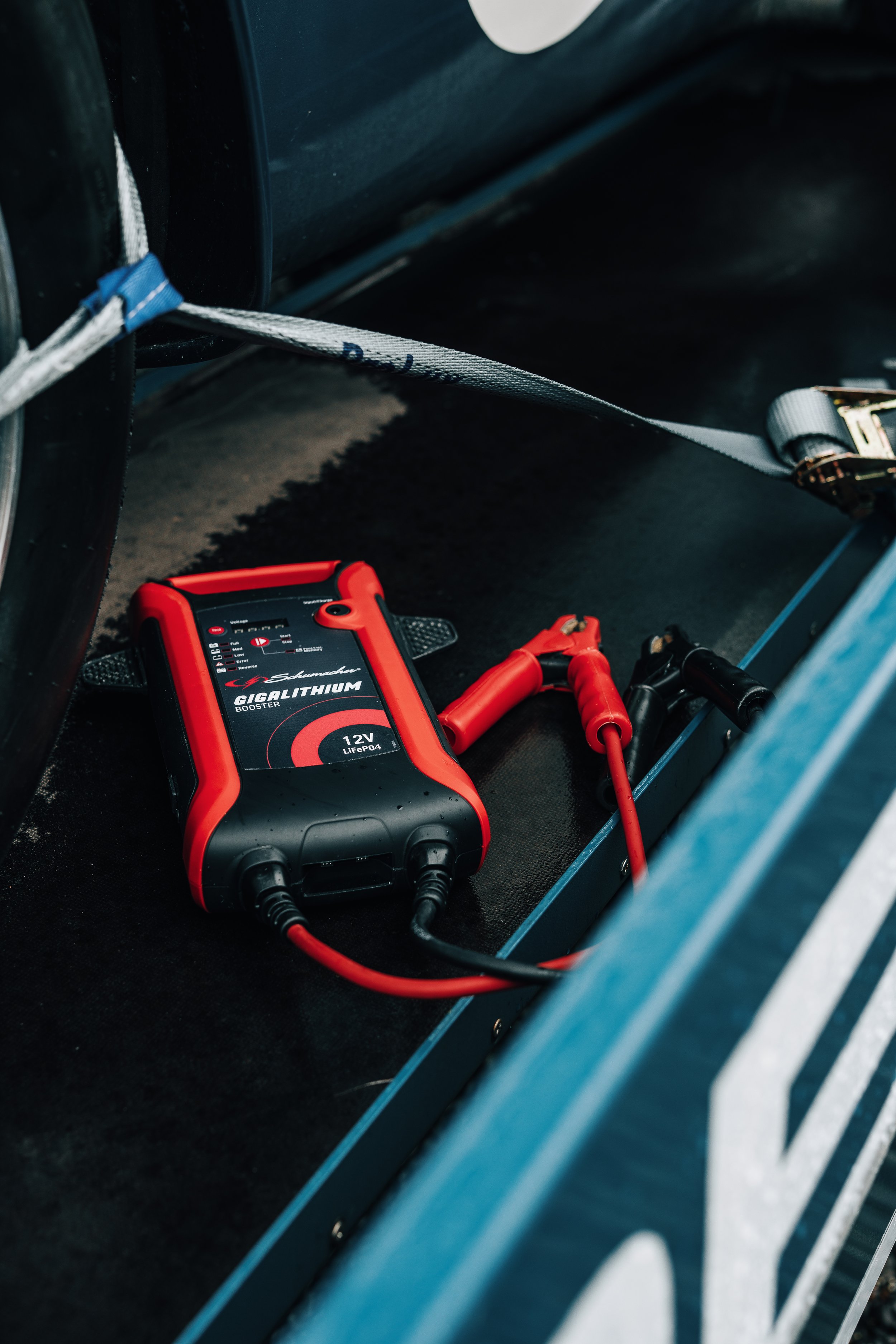 A red Schumacher Giga Lithium booster battery charger connected to black and red cables inside a vehicle's compartment with a blue frame.