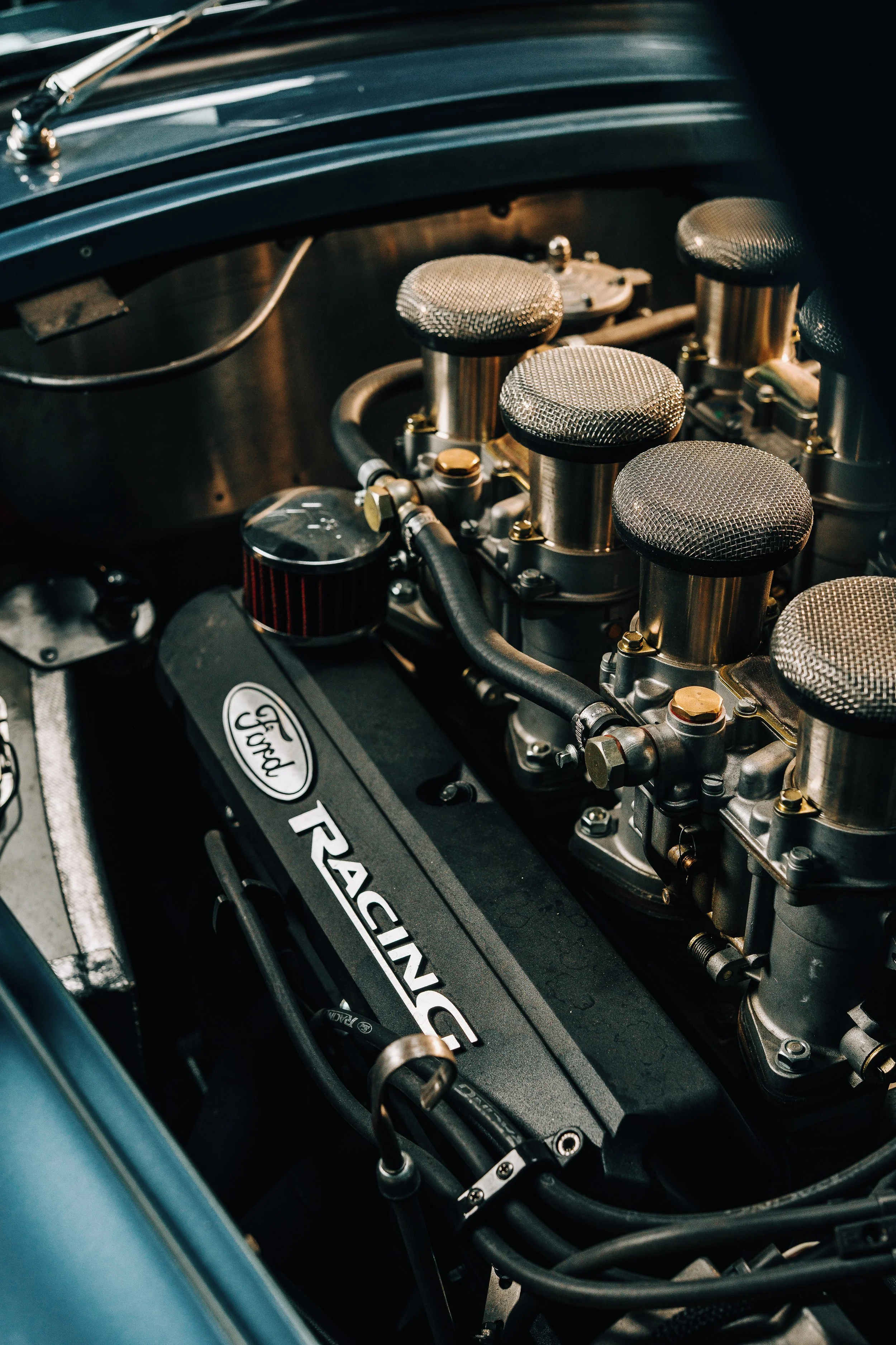 Close-up view of a classic car engine with multiple metallic intake stacks, a black valve cover with the Ford logo, and various hoses and fittings.