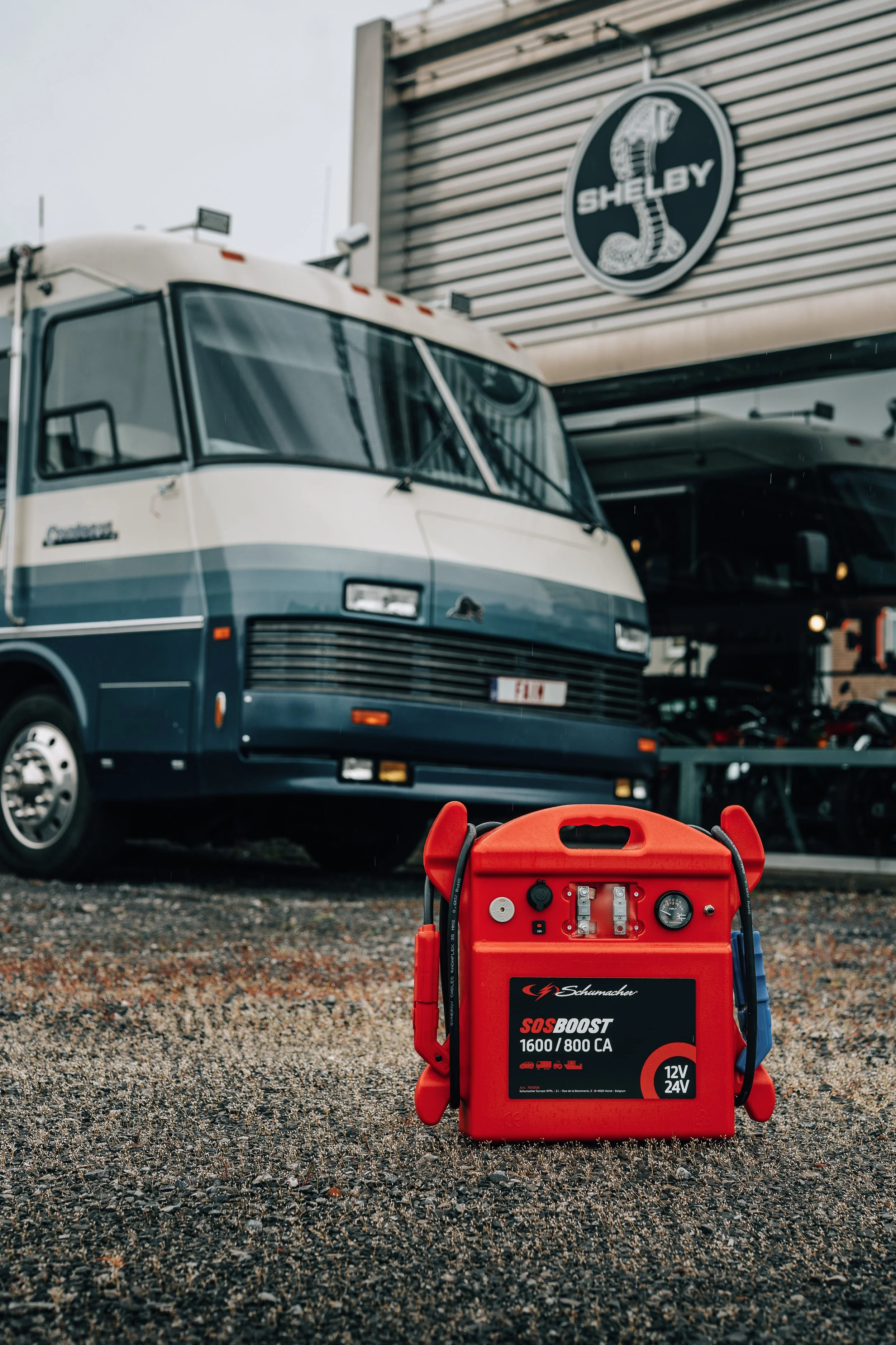 A red Schumacher SOS Boost car jump starter with 1600/800 CA rating and 12V/24V capacity, placed on gravel in front of a parked blue and white motorhome with a Shelby logo on the building behind.
