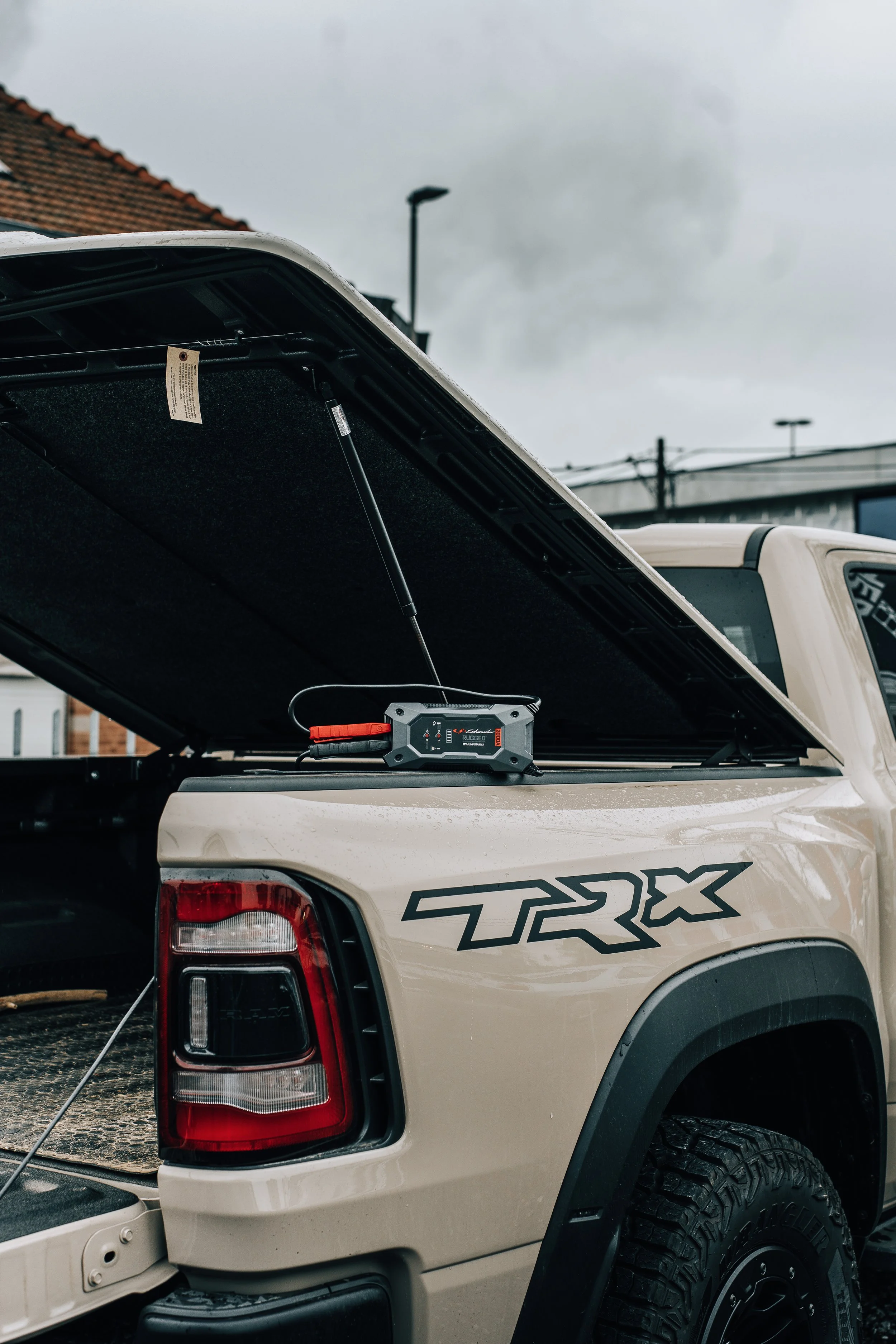 The back of a beige Toyota TRX pickup truck with an open tailgate, showing a portable jump starter device on the truck bed, with a cloudy sky in the background and part of a house roof visible.