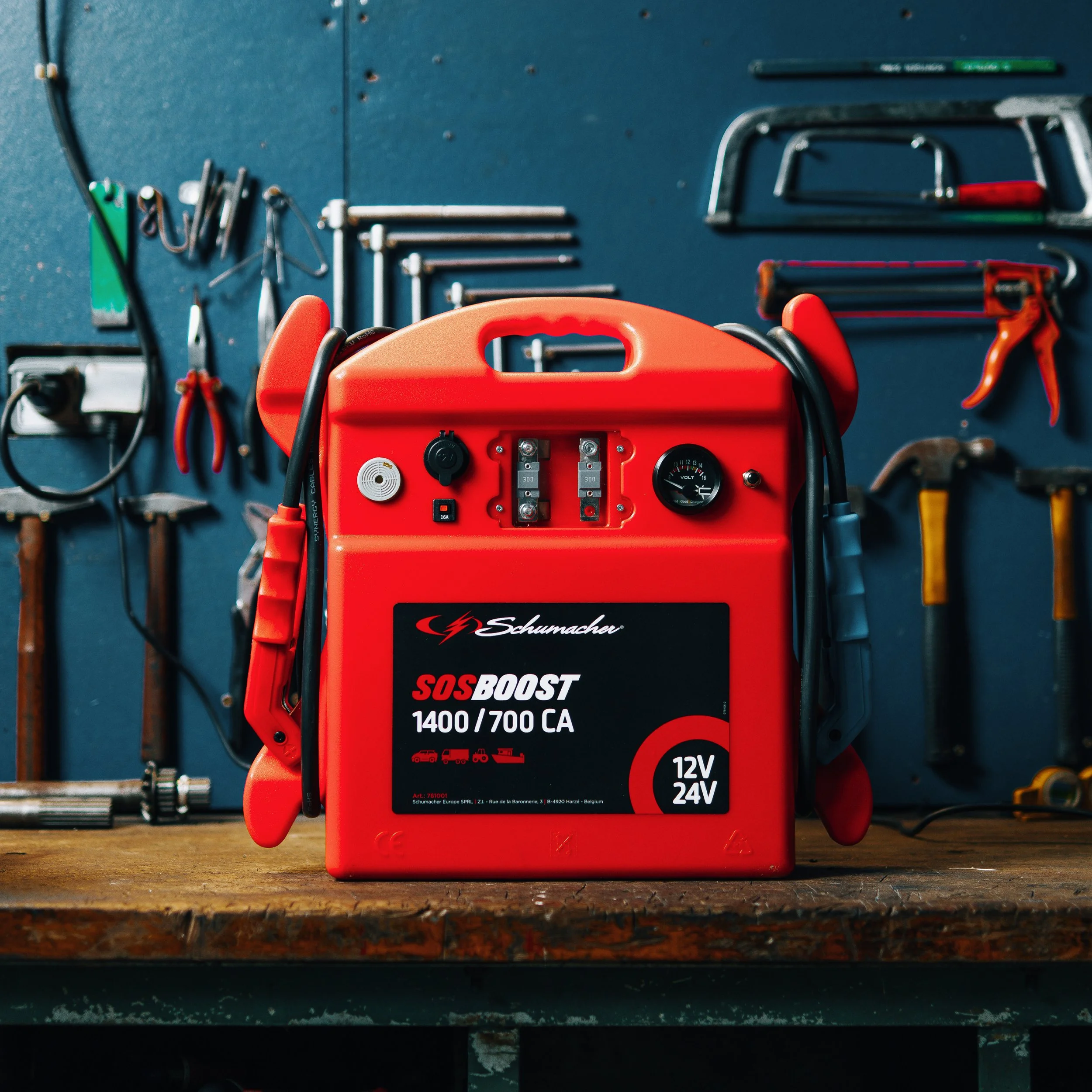 A red Schumacher automotive booster with black and red controls, a voltmeter, and a power switch on a wooden workbench with tools in the background.