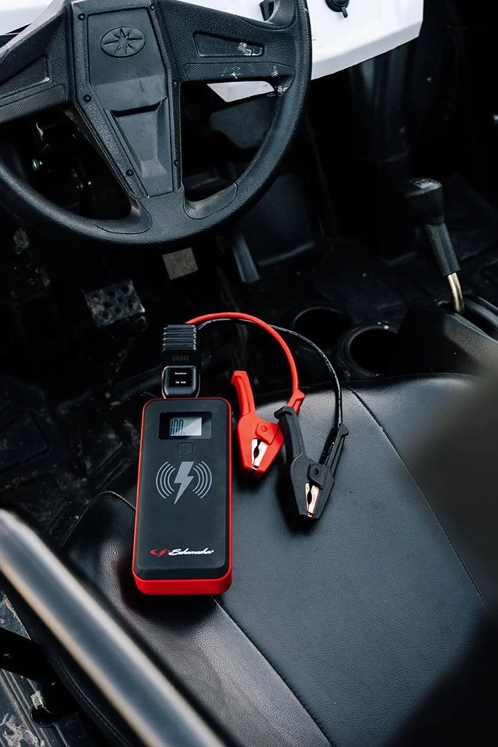 A diagnostic device with wireless charging symbol, connected to alligator clips, placed on a black vehicle seat inside a vehicle.