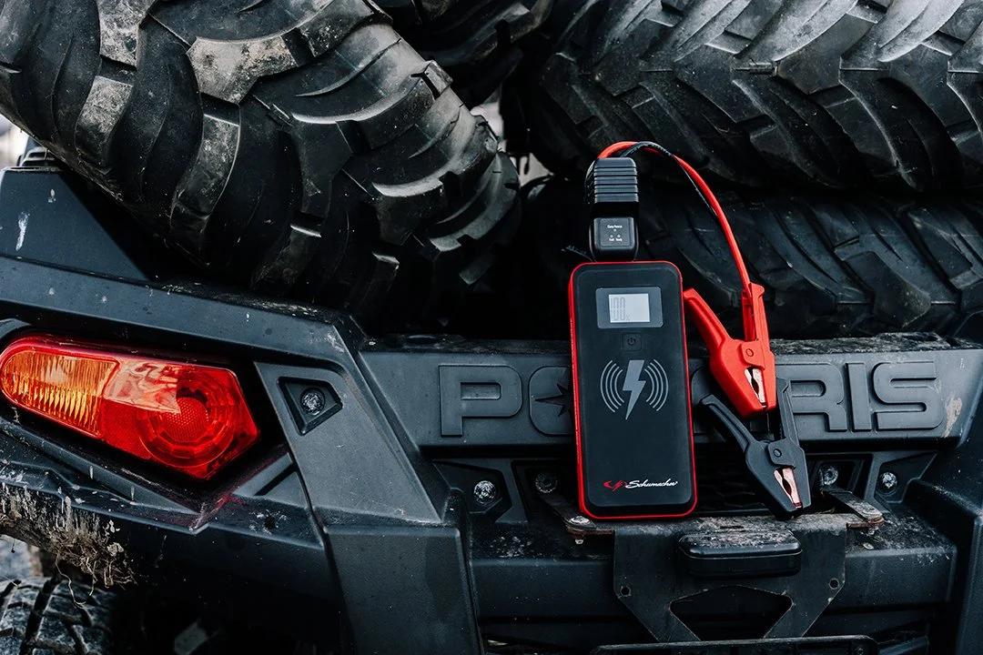 A battery jump starter connected to a Polaris off-road vehicle's battery, with jumper cables attached, parked on muddy ground.