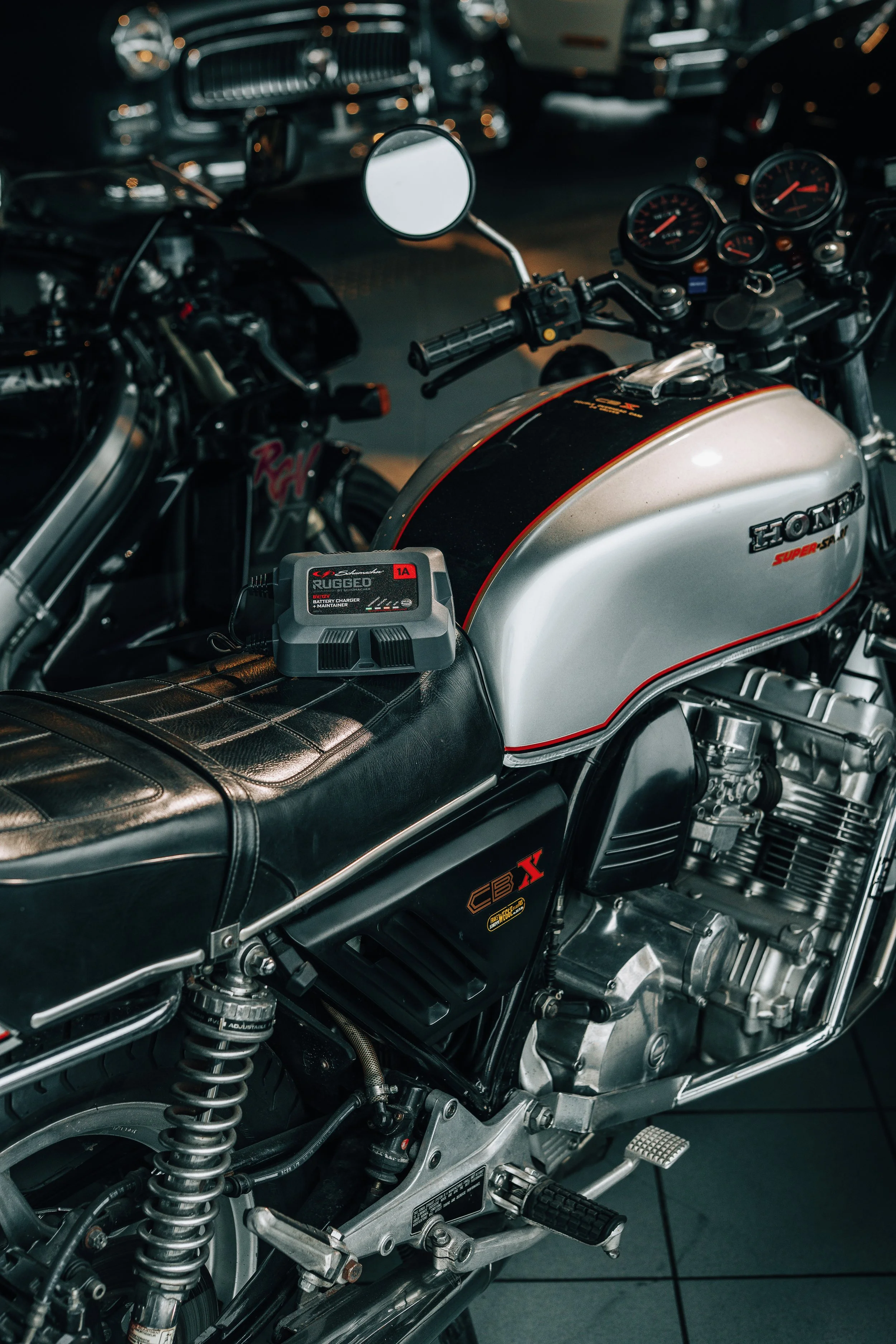 Close-up of a Honda motorcycle with a silver fuel tank and black seat, showing the handlebar, gauges, and suspension details in a dimly lit garage.