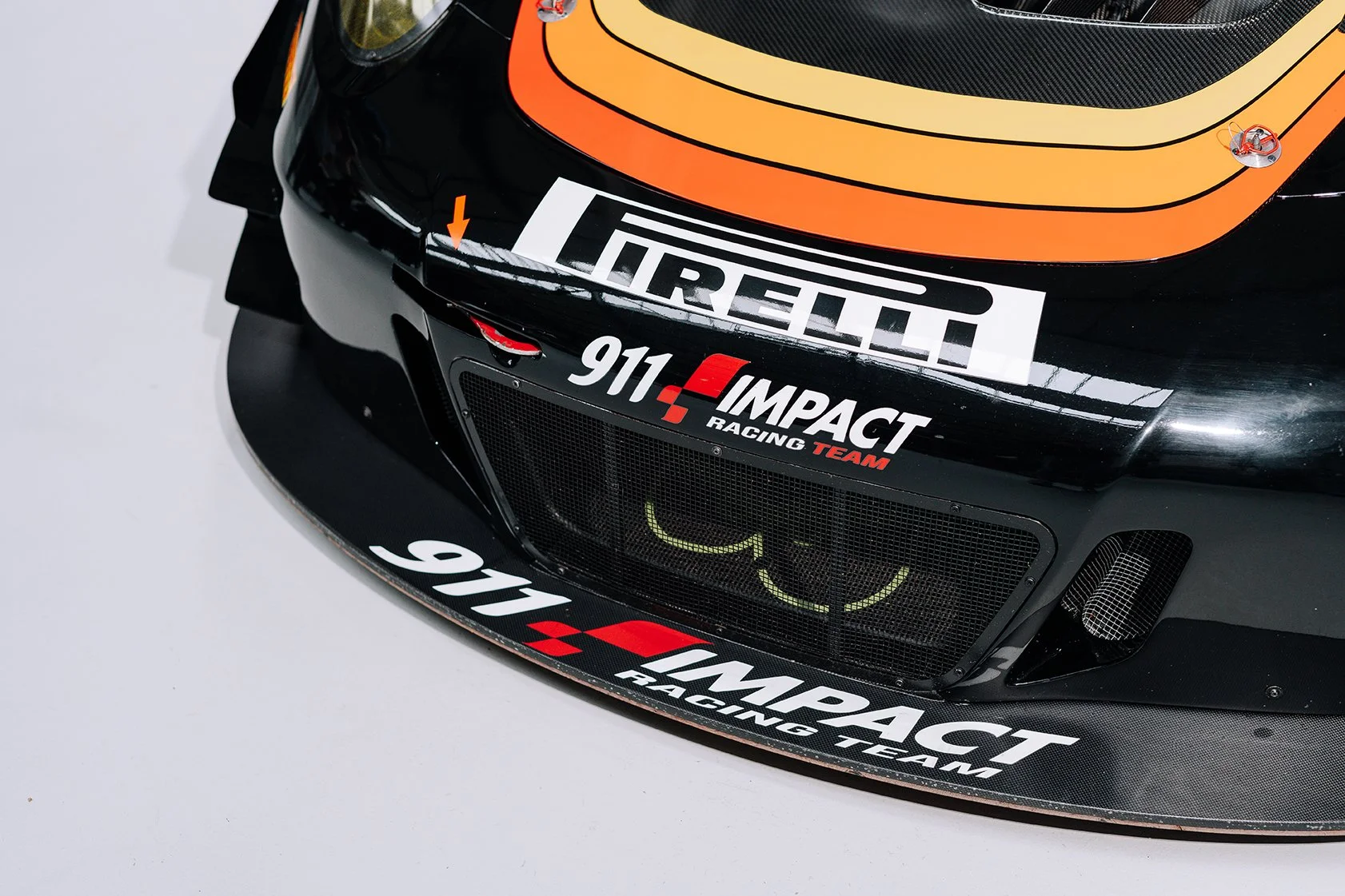 Close-up of the front of a black racing car with sponsor logos including Pirelli, Impact Racing, and Team 911 Impacts, featuring yellow and orange accents and a mesh grille.