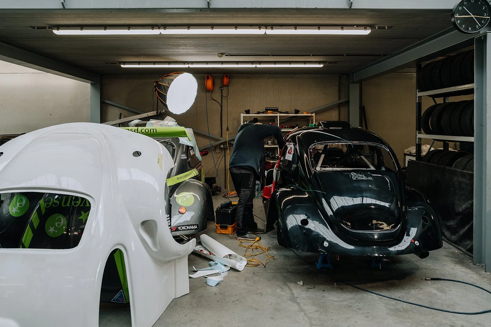 Inside a garage or workshop with two racing car bodies, one white and one black, being worked on by a person. Shelves with tires are visible, along with tools and lighting.