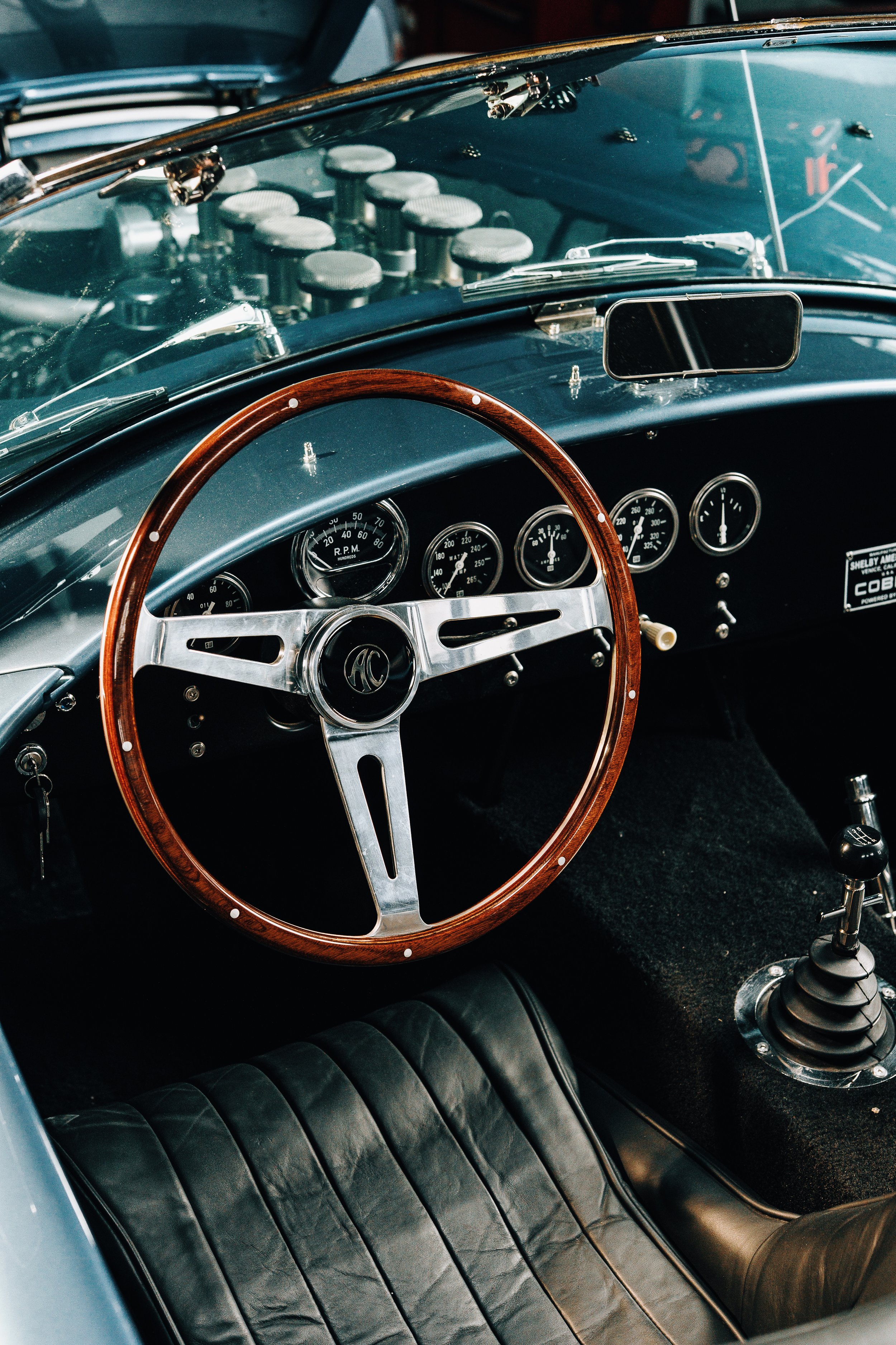 Interior of a vintage car showing a black leather seat, a wooden steering wheel, and a dashboard with multiple gauges, a mirror, and a box of engine components visible through the windshield.