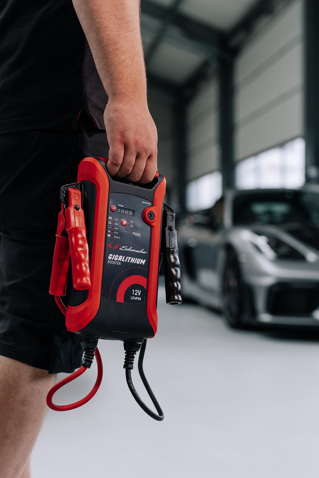 A person holding a Schumacher Gigalithium booster with red and black design, in an indoor garage with cars in the background.