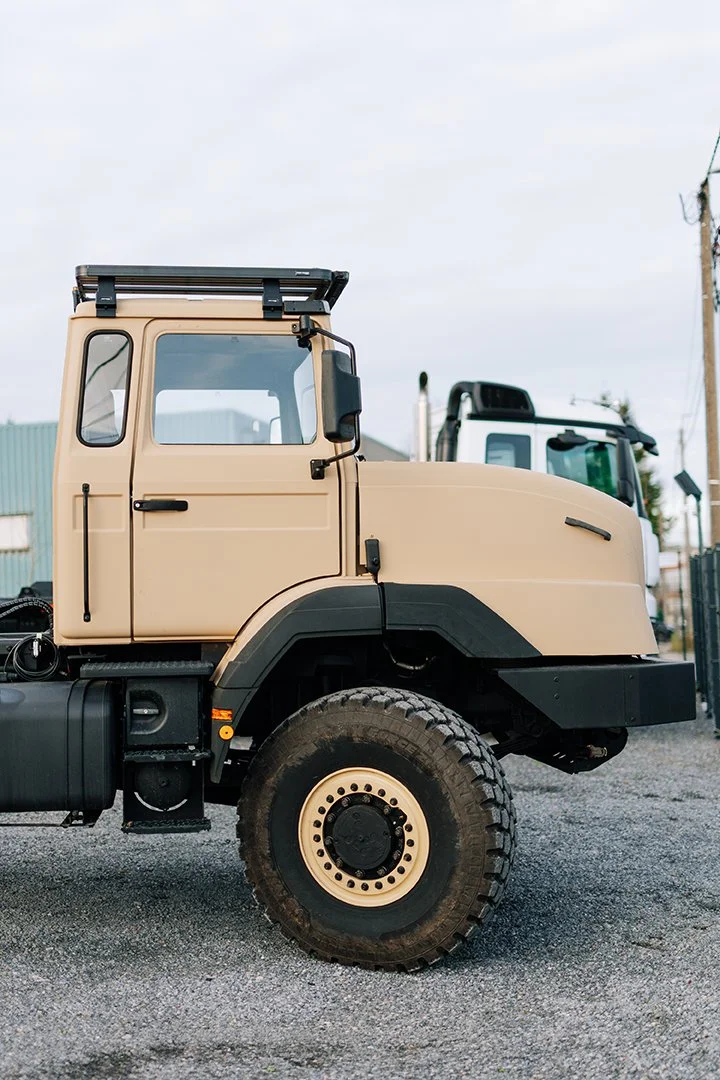 A beige industrial truck parked on a gravel lot with another truck in the background under an overcast sky.