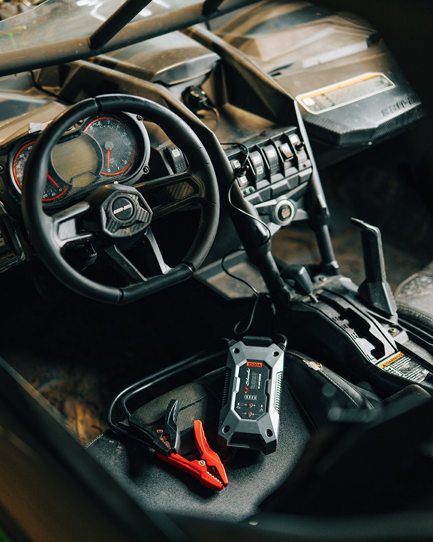Inside a rugged off-road vehicle with a steering wheel, dashboard, and center console, featuring a portable jump starter and jumper cables lying on the seat.