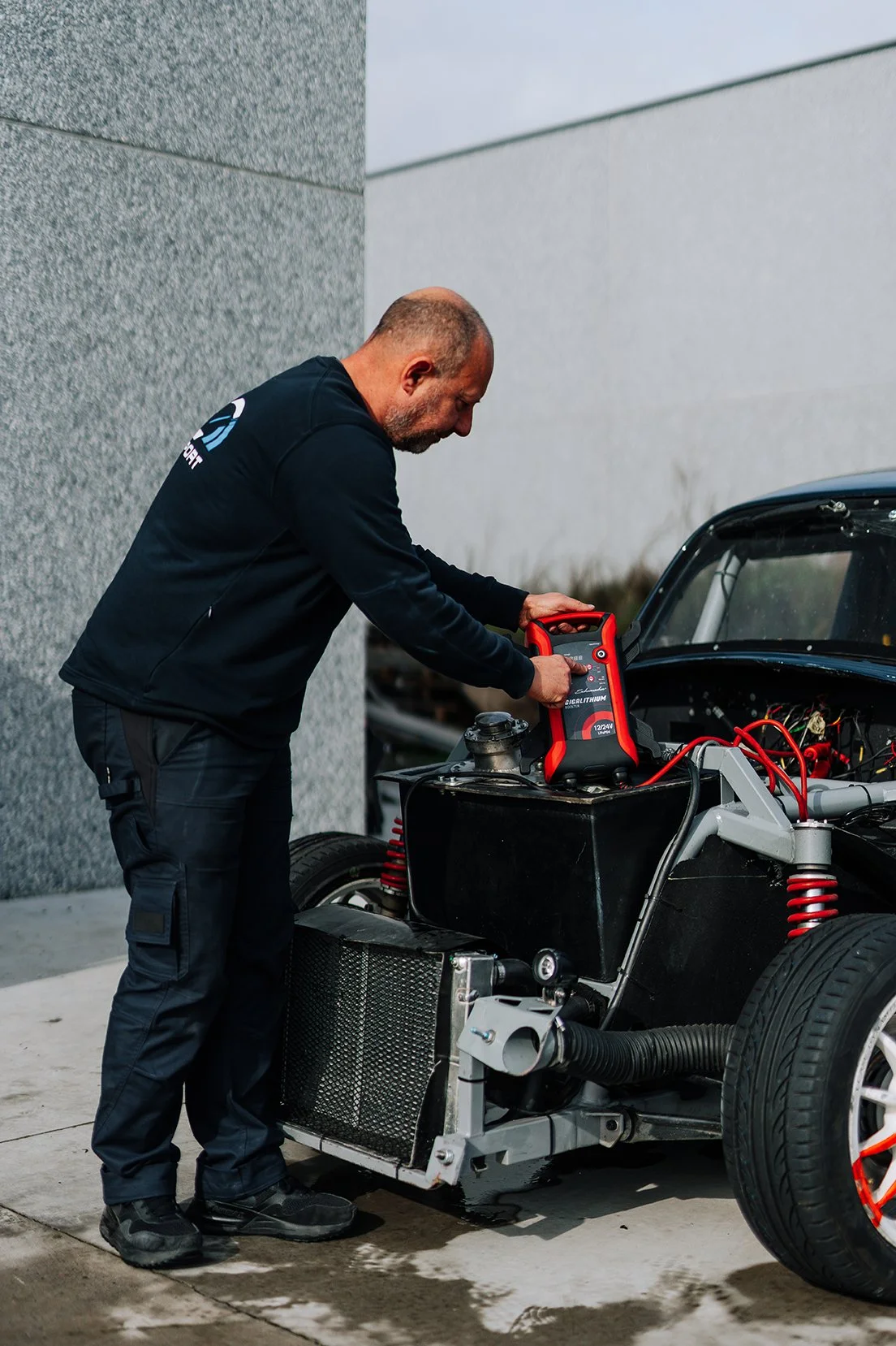 A man inspecting or working on a race car with an electronic device connected to the engine, outdoors near a gray building.