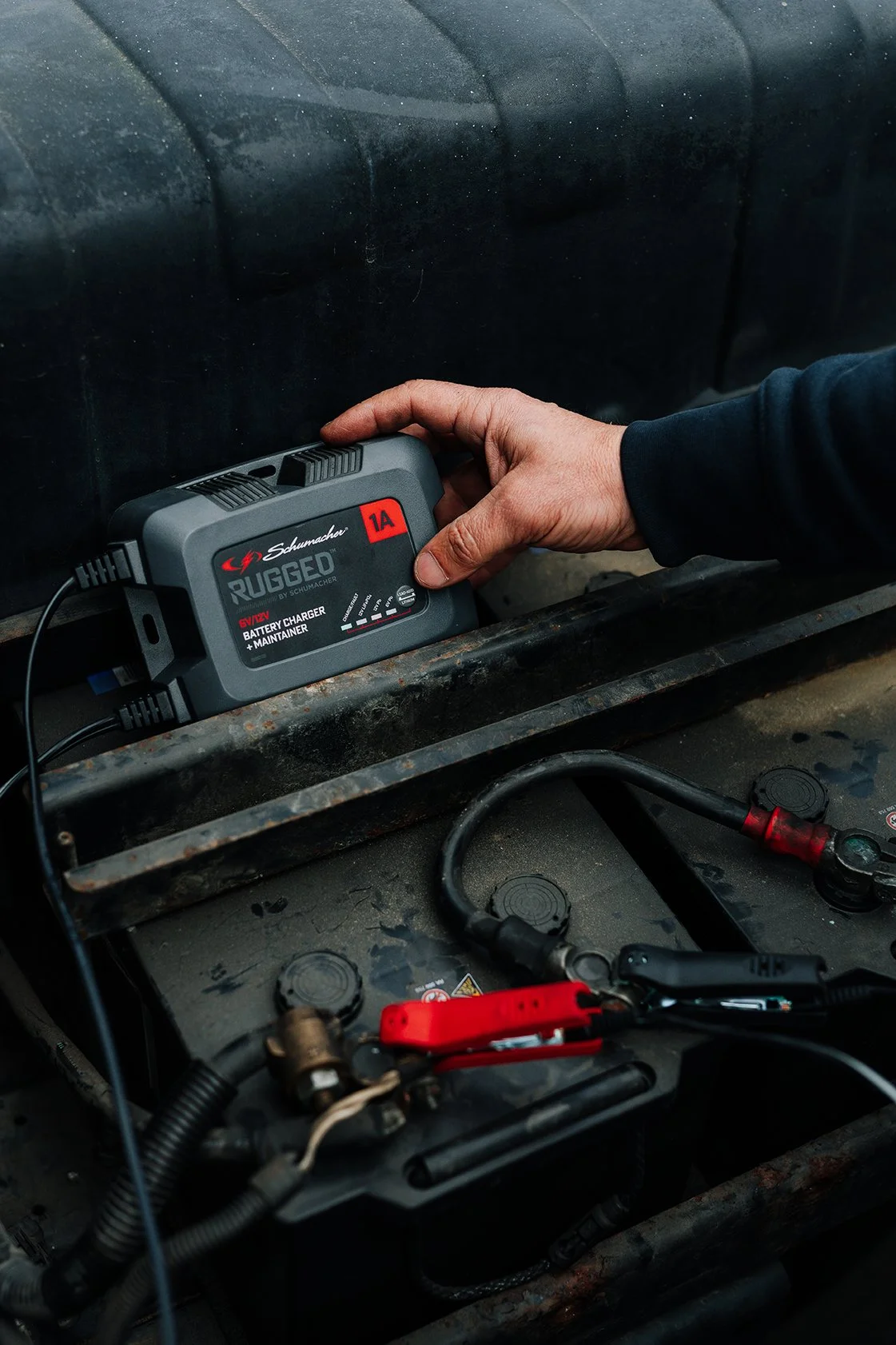 A person holding a Schumacher Rugged 1A battery charger and maintainer connected to a vehicle battery in a vehicle's engine compartment.