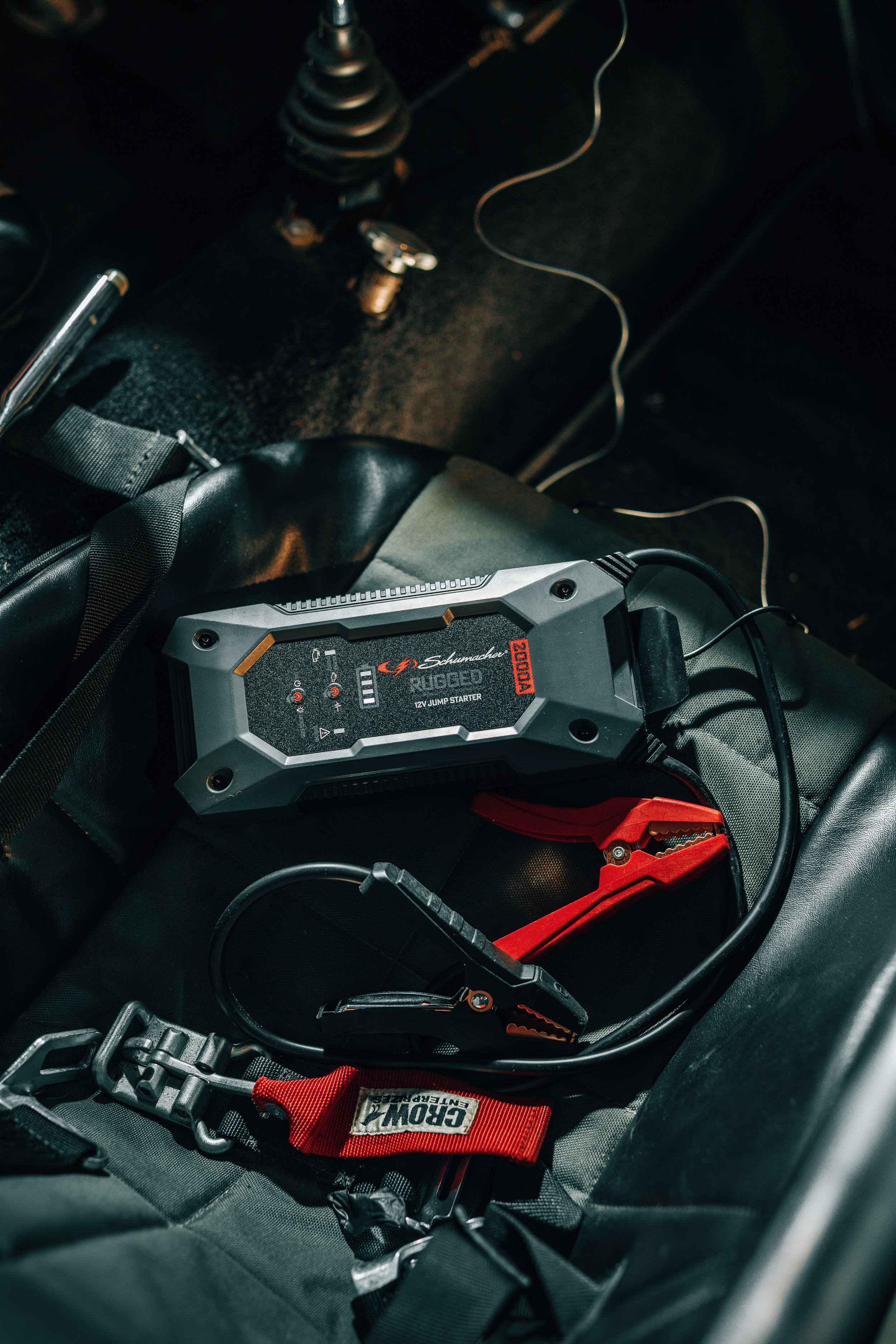A close-up of a car interior showing a jump starter device with red and black alligator clips connected to the car's battery. The jump starter is labeled 'Schumacher' and 'RB1680.' There are various wires, a seatbelt, and parts of the car's interior 