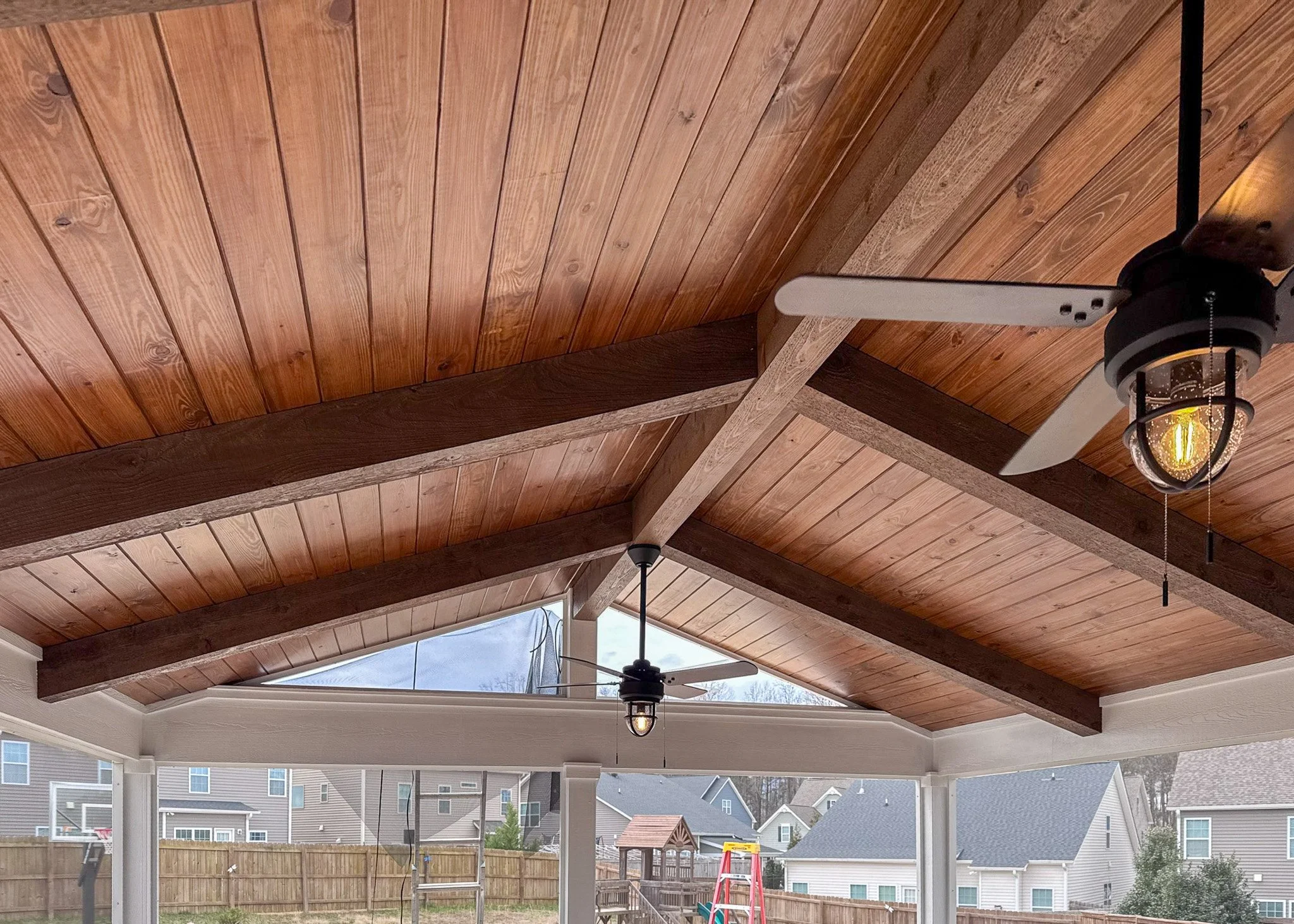 Photo of a porch with a wooden ceiling, ceiling fans, exposed beams, and view of a backyard with a ladder, slide, and a person wearing a hat.