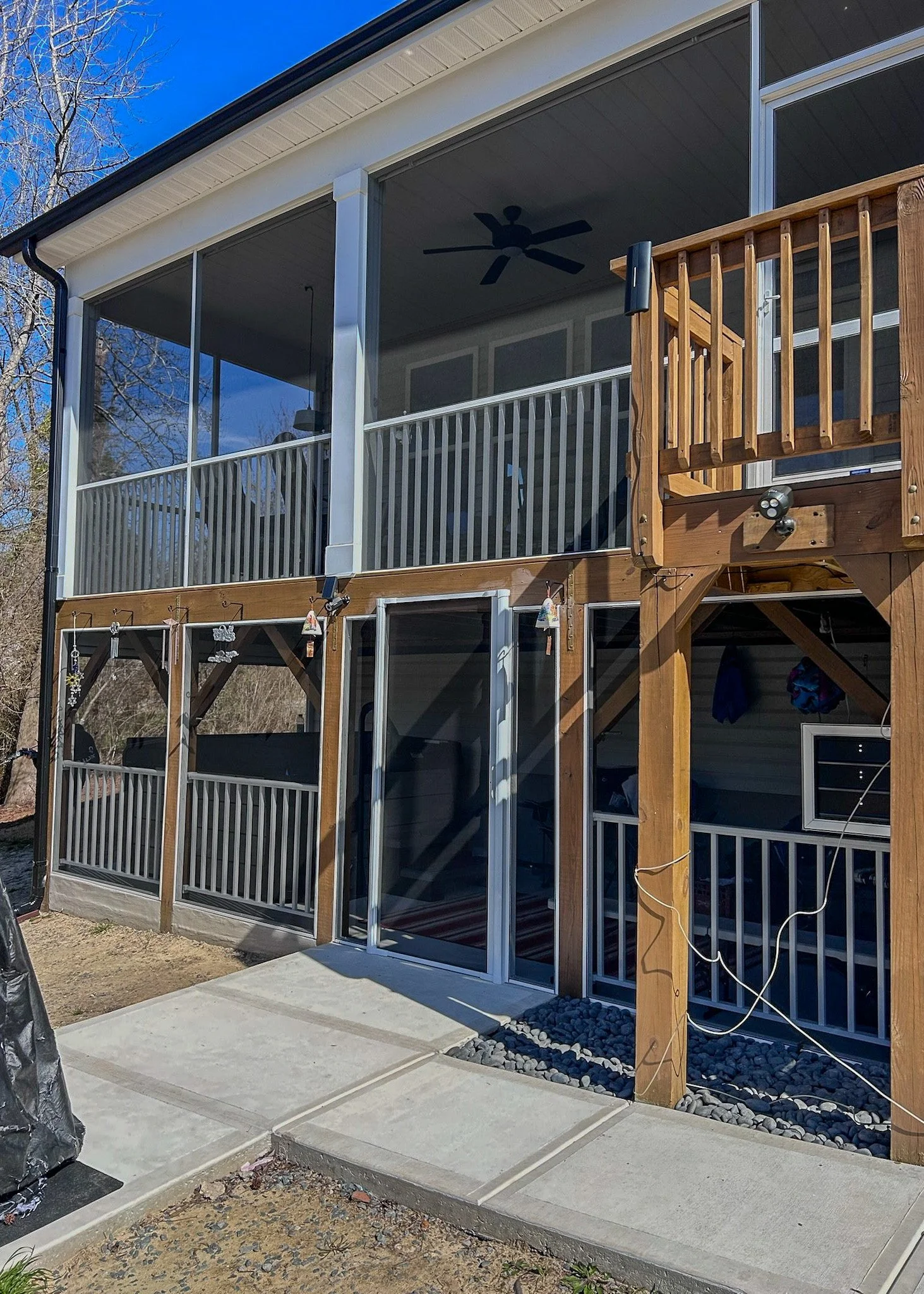 Newly constructed two-story house with a covered porch, screen enclosed ground level, open upper level with railings, wooden staircase, and a concrete patio in the backyard.