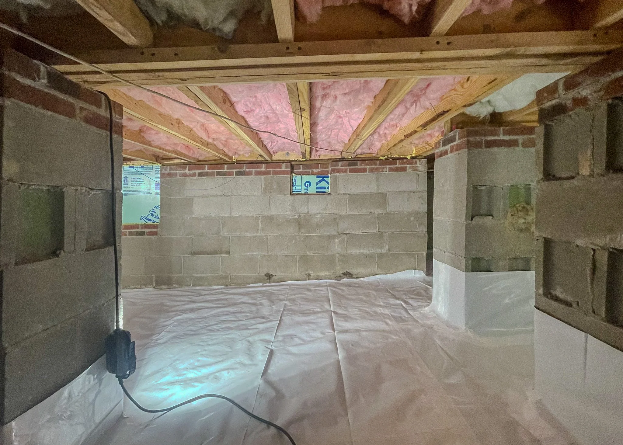 Interior view of a building under construction or renovation, showing exposed brick and concrete walls, insulation pink fiberglass on the ceiling, and wooden beams, with a plastic sheet covering the floor.