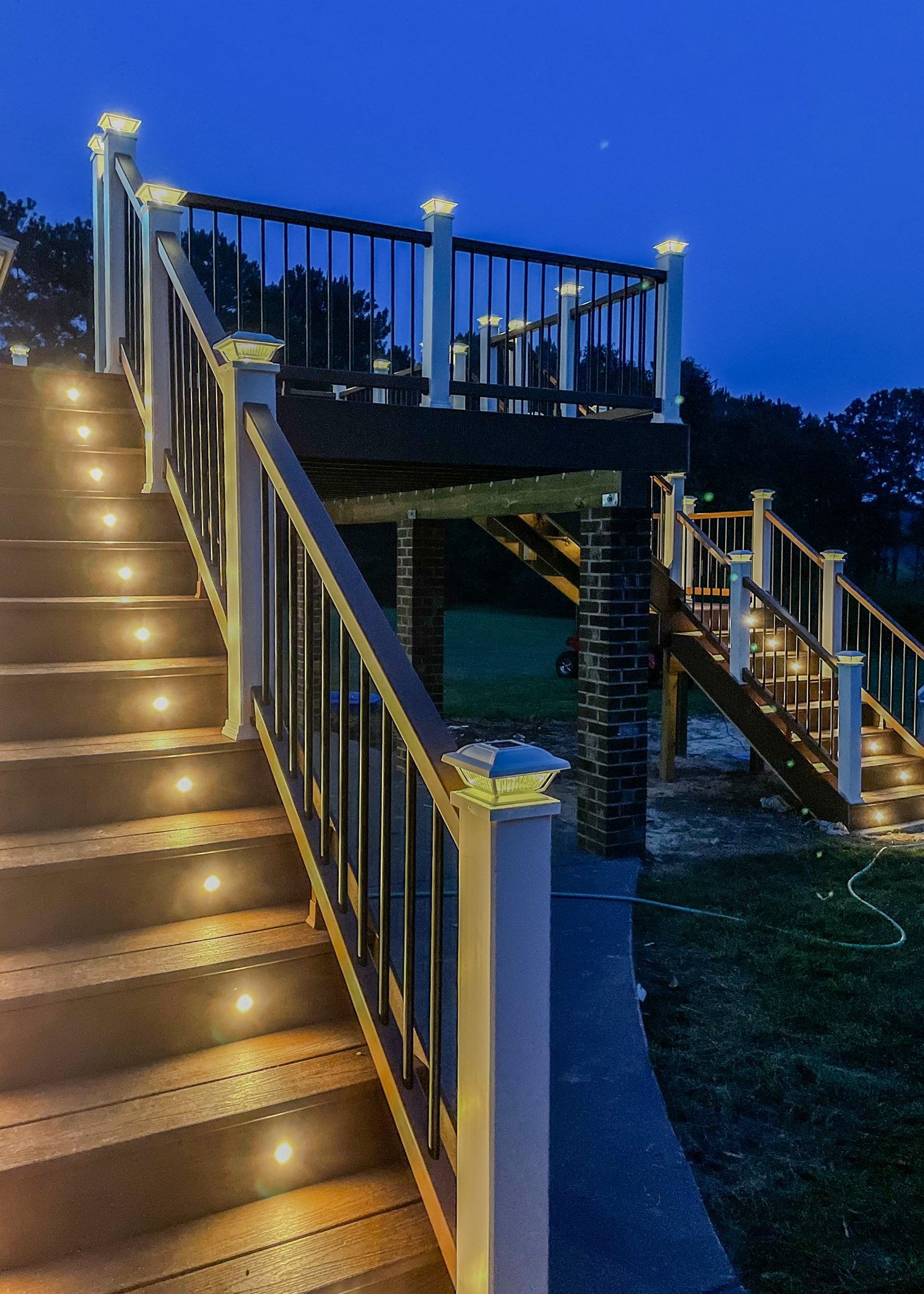 Outdoor staircase with black and white railing, illuminated by built-in lights, at dusk with trees and dark blue sky in the background.