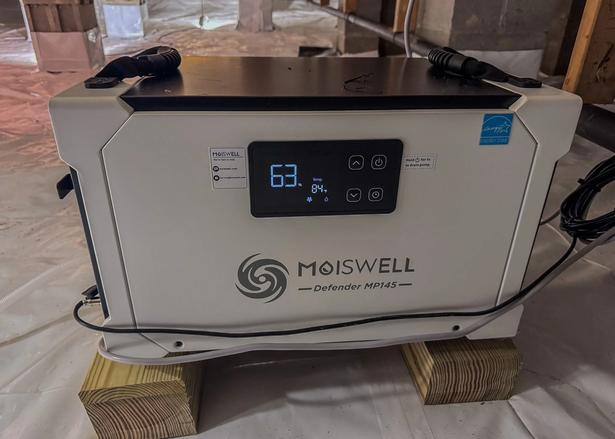 A large white moisture control machine with a digital display showing the number 63, placed on two wooden blocks on a construction site with unfinished walls.