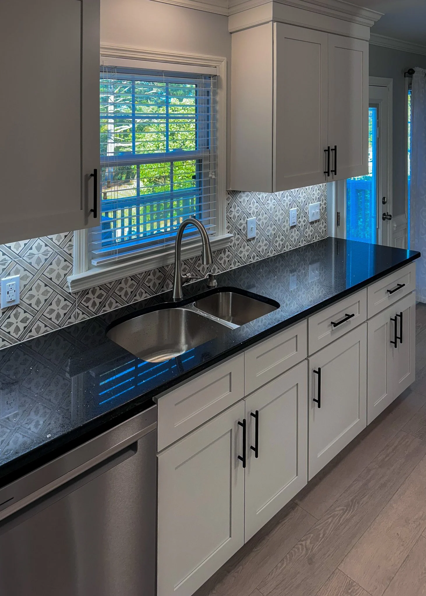 Kitchen with white cabinets, black granite countertop, backsplash with geometric tile pattern, double basin stainless steel sink, window with blinds, and light-colored flooring.