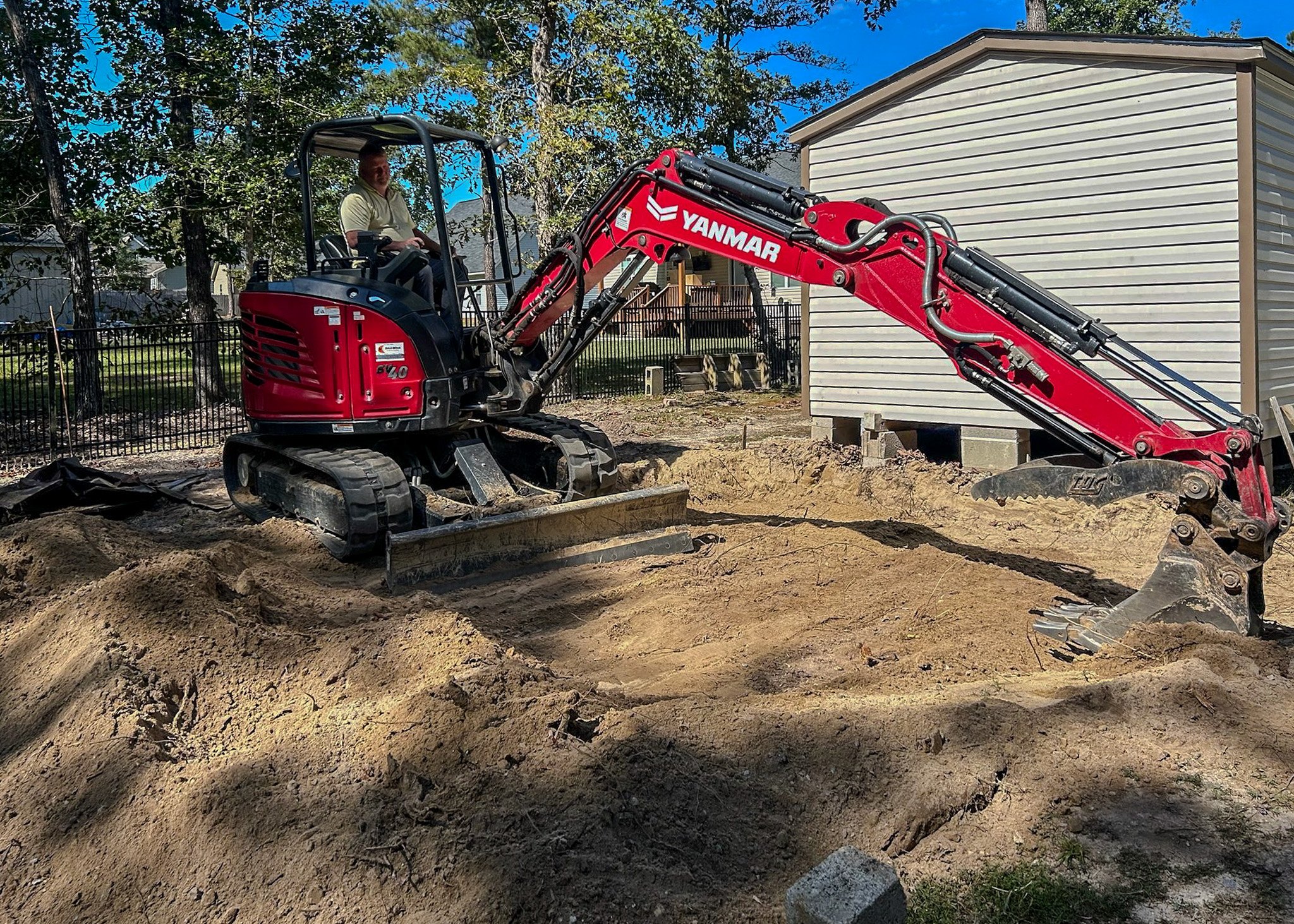 A person operating a red Yanmar mini excavator digging in a residential backyard, with trees and a house in the background.