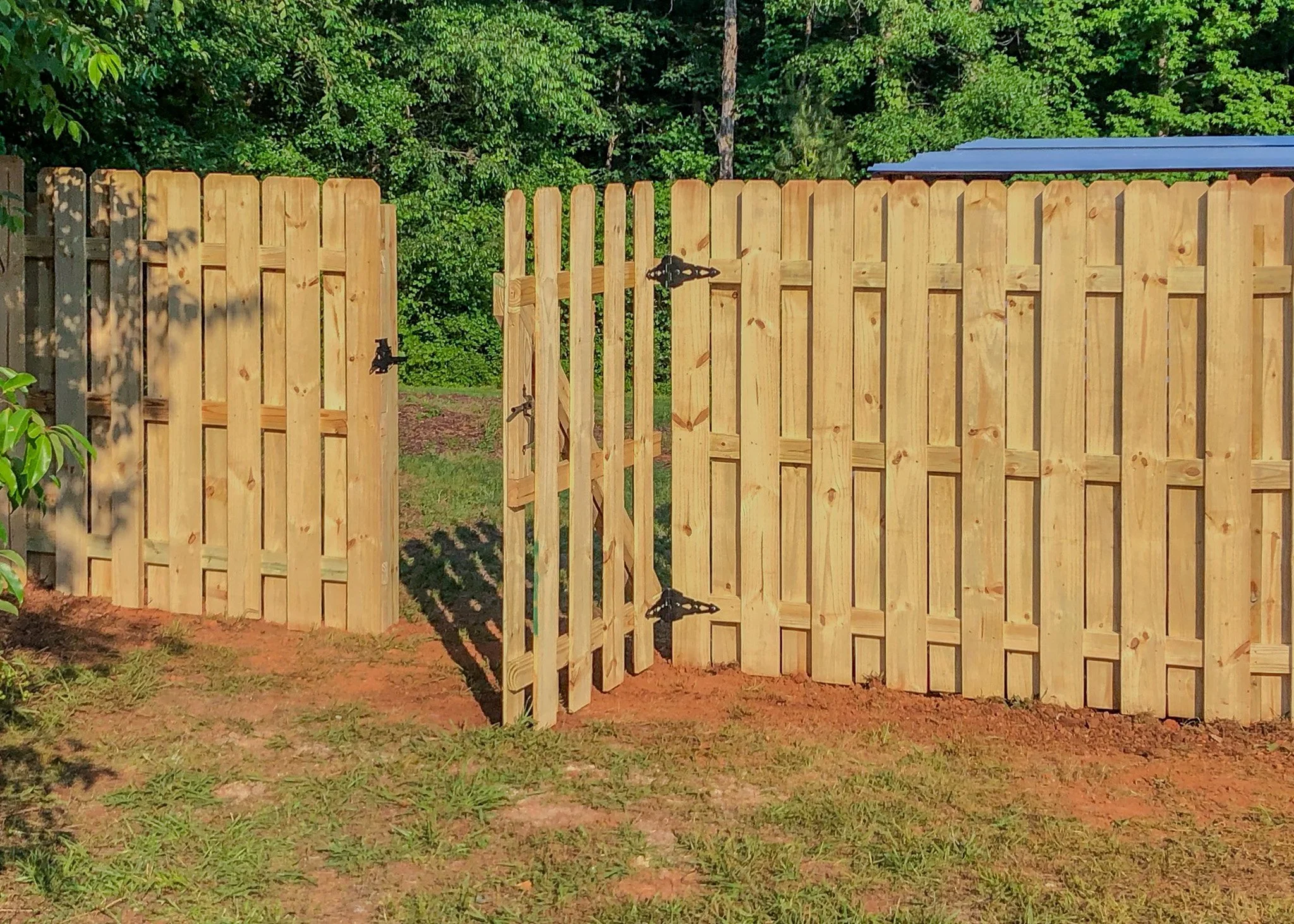A wooden gate with a smaller gate in the middle, all made of light-colored vertical wooden planks, set against a background of green foliage and trees.