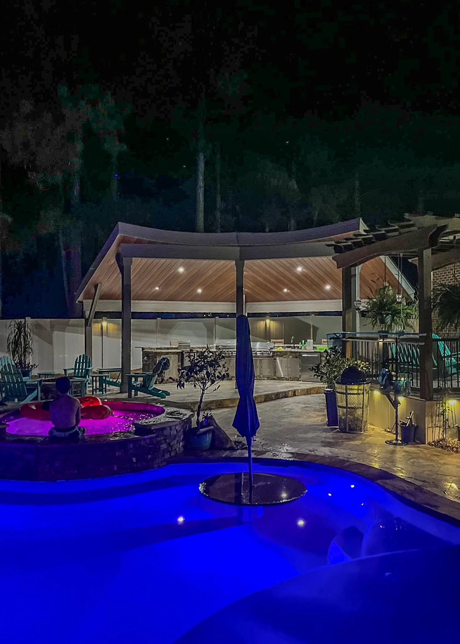 Night view of a backyard pool area illuminated with colorful lights, featuring a person sitting in a hot tub, outdoor seating, a shaded pavilion, and a bar area in the background.