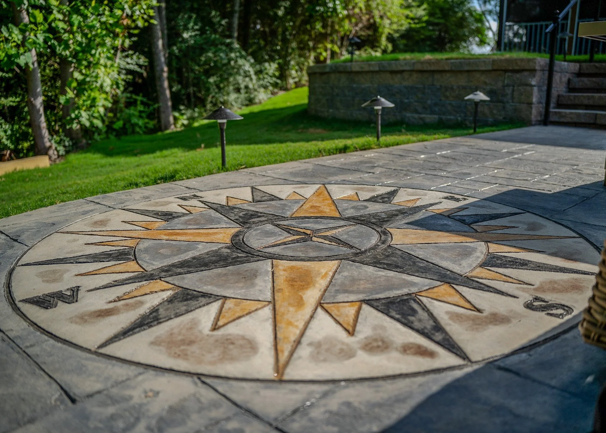 A decorative mosaic compass rose on an outdoor patio with grass, trees, and outdoor lighting fixtures in the background.