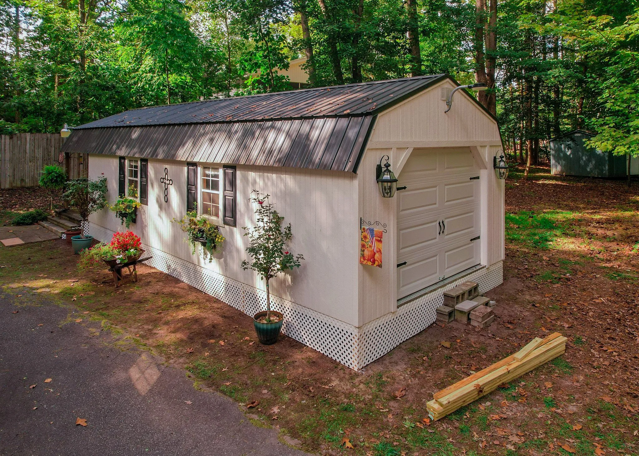 A small white garage with black shutters, a metal roof, and a decorative cross on the wall, surrounded by potted plants and trees in a backyard.