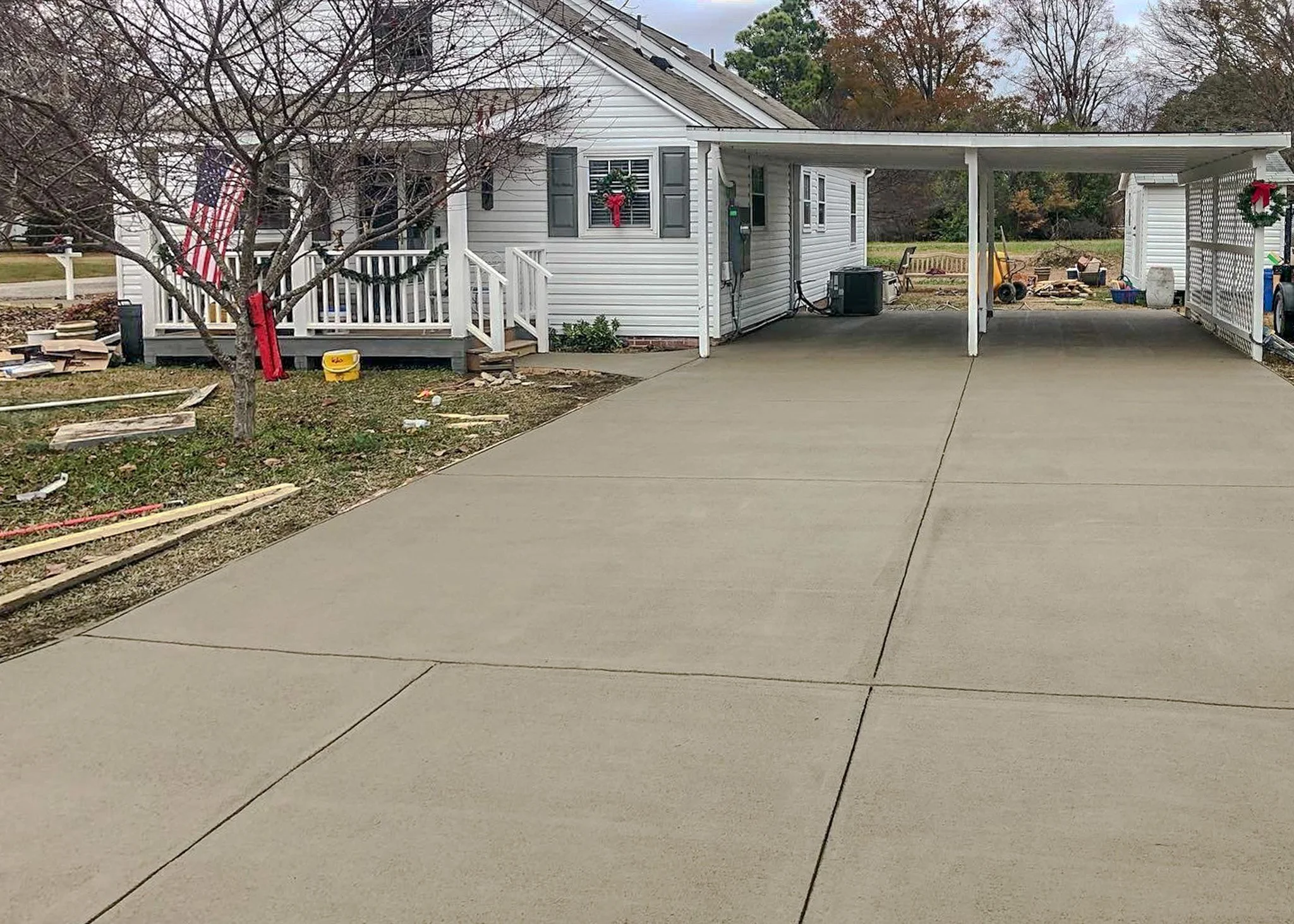Newly poured concrete driveway leading to a white house with a porch decorated for Christmas, a leafless tree on the left, and a shed on the right with construction tools and scattered debris.