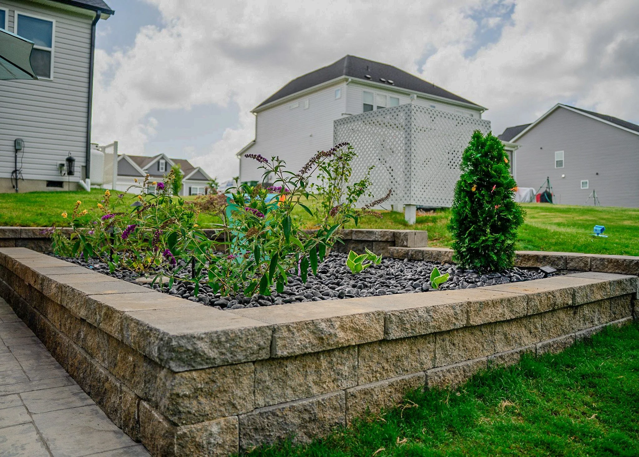 A backyard garden with a raised flower bed made of stacked stone, filled with plants and black decorative rocks, with a backdrop of houses and a partly cloudy sky.