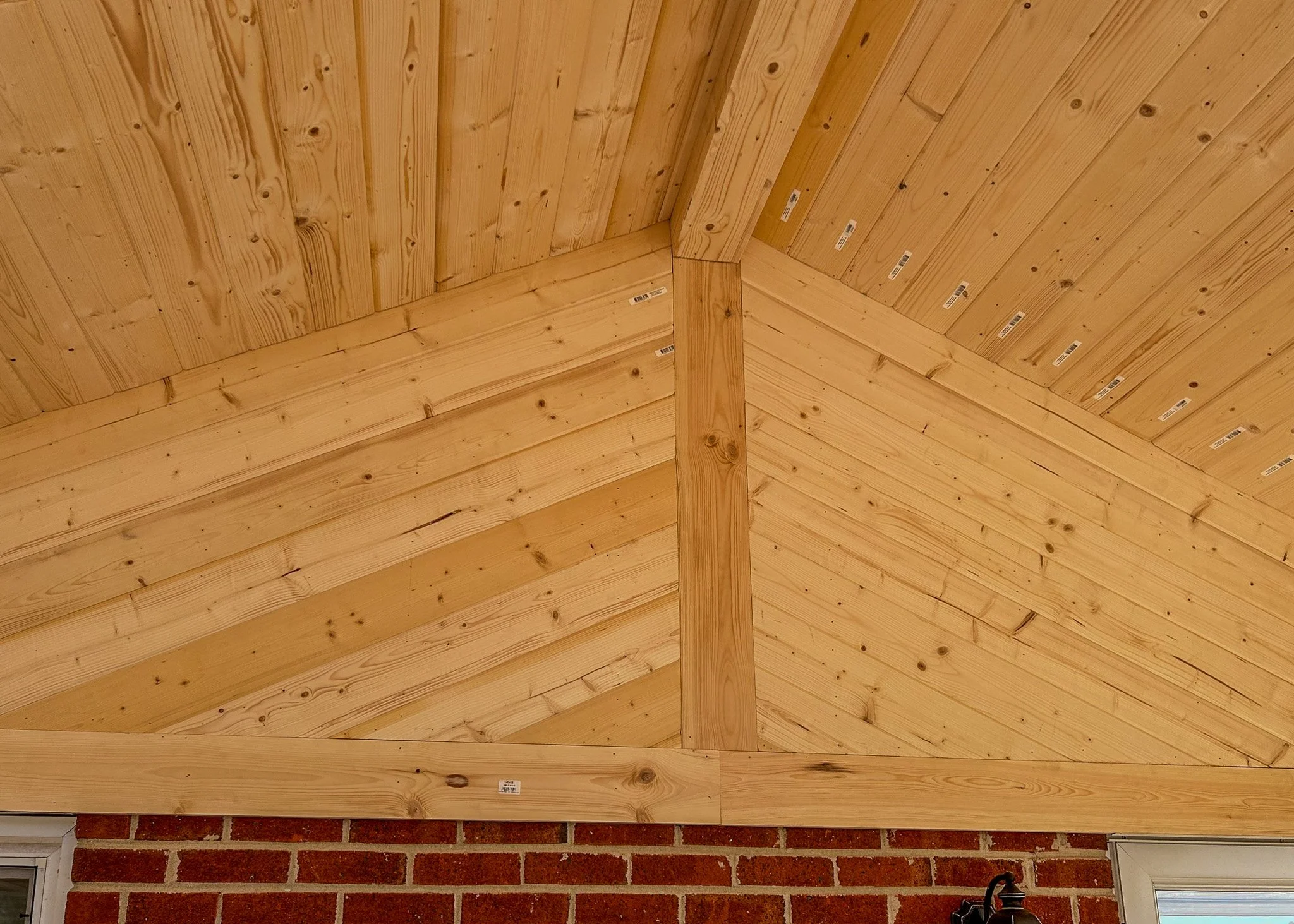 Interior view of a ceiling with exposed wooden beams and tongue-and-groove wooden paneling, above a brick wall with a window, outdoor light fixture, and ladder.