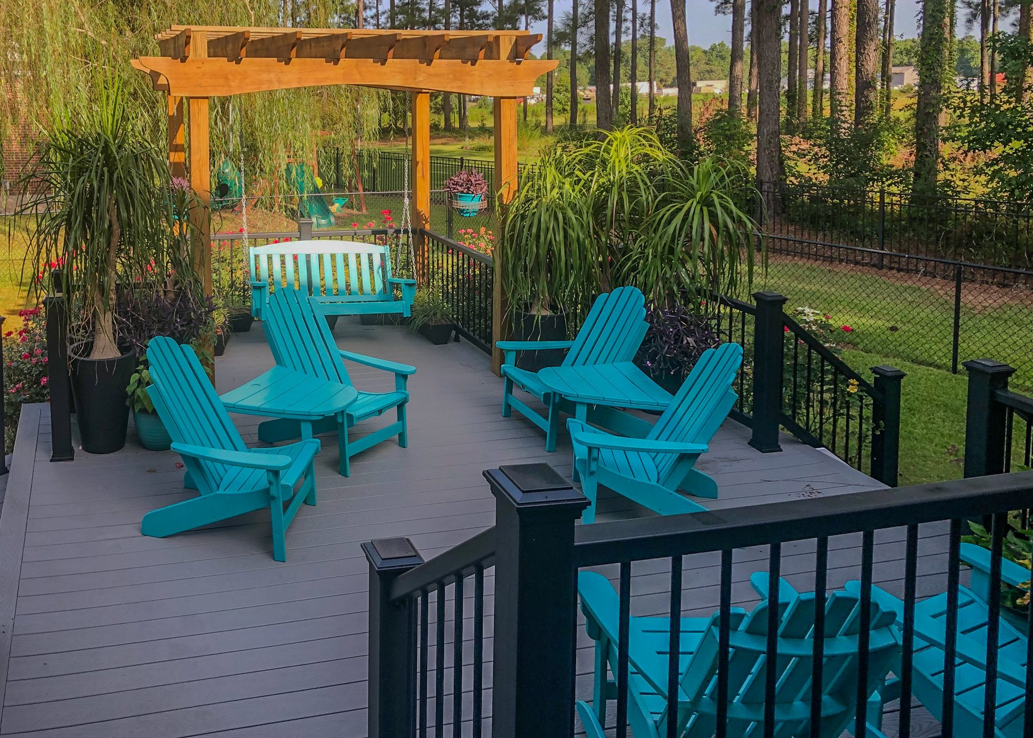 A backyard patio with four turquoise Adirondack chairs arranged in pairs facing each other, surrounded by potted plants and greenery, with a wooden swing under a pergola in the background.