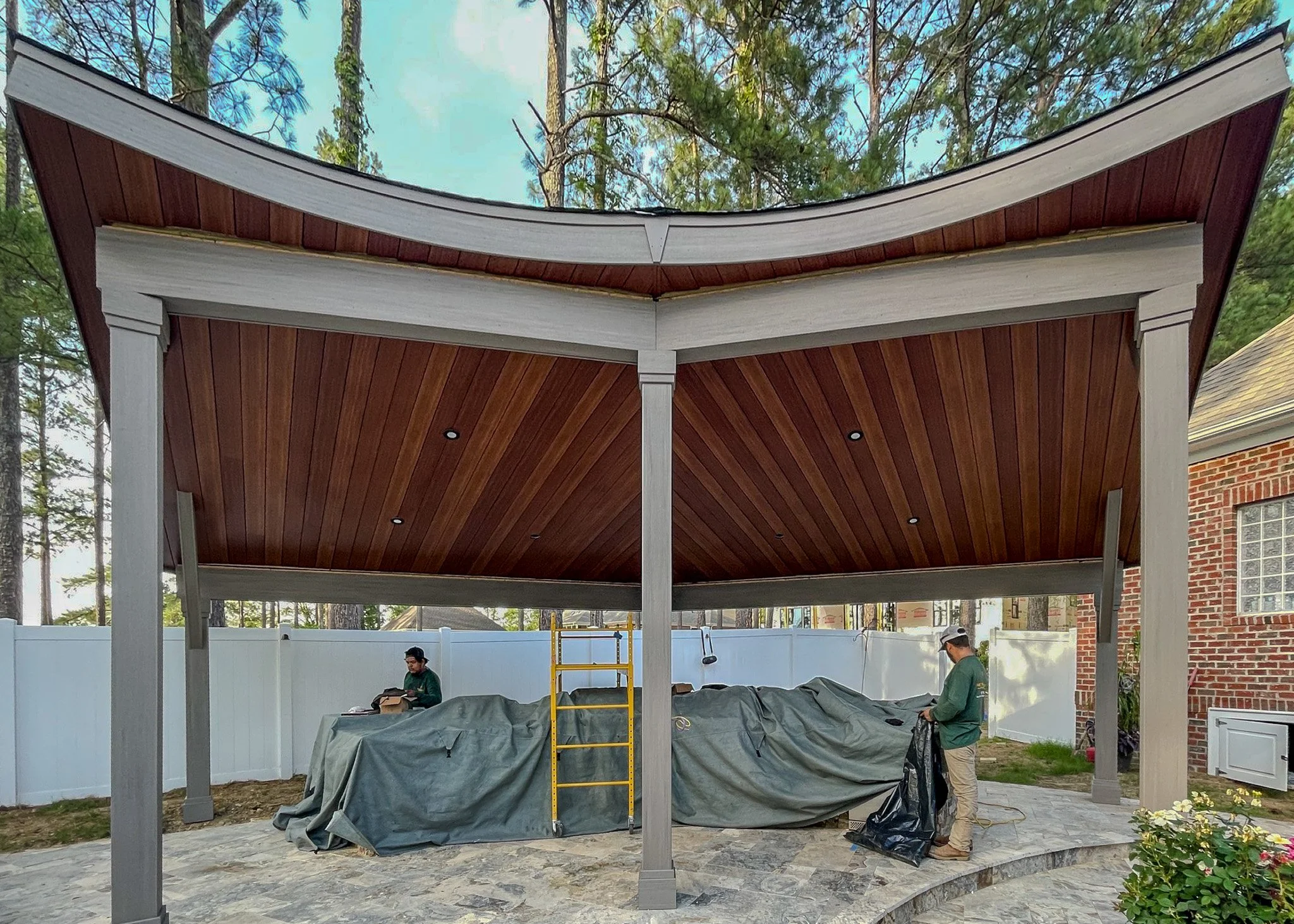 Two workers setting up a covered outdoor patio or gazebo extension with a wooden ceiling over a stone patio, nearby a red brick house and a white fence surrounded by trees.