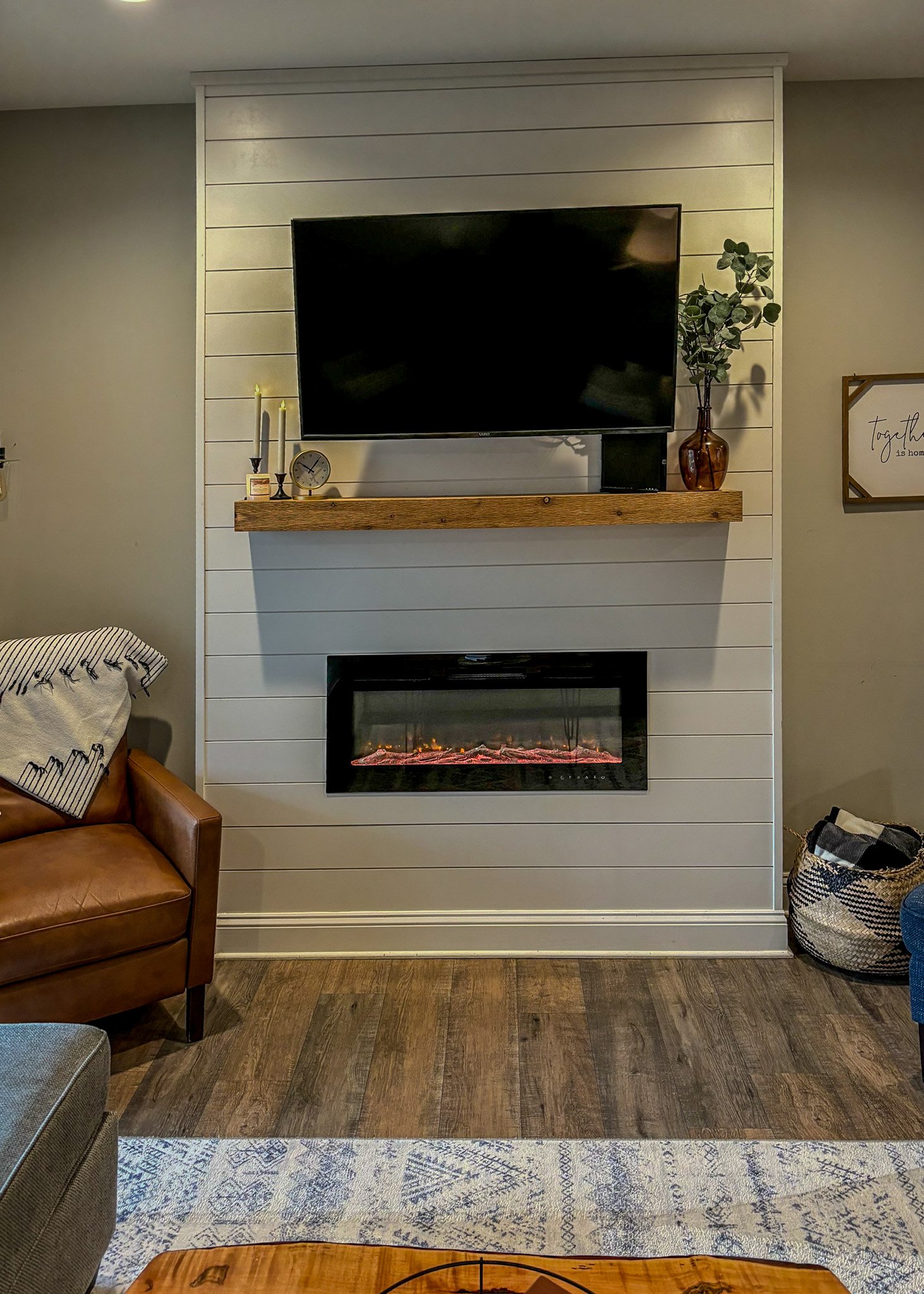 Living room with a wall-mounted TV above a fireplace, a wooden mantel with candles, a clock, and a plant in a brown vase. Furniture includes a brown armchair with a blanket, a gray sofa, a wooden coffee table with books, and a blue chair. There is a 