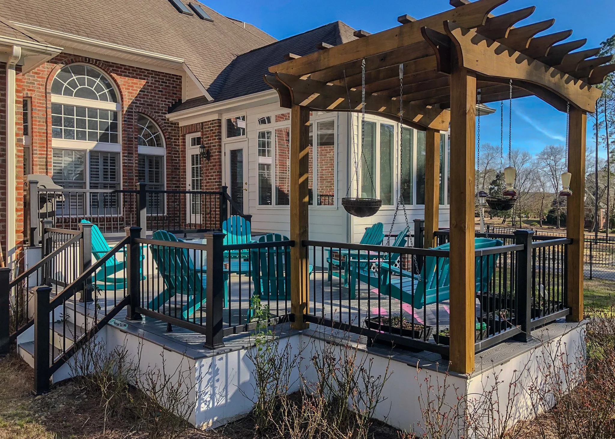 A backyard patio with turquoise Adirondack chairs under a wooden pergola, attached to a brick house with white-framed windows and a white enclosed porch.