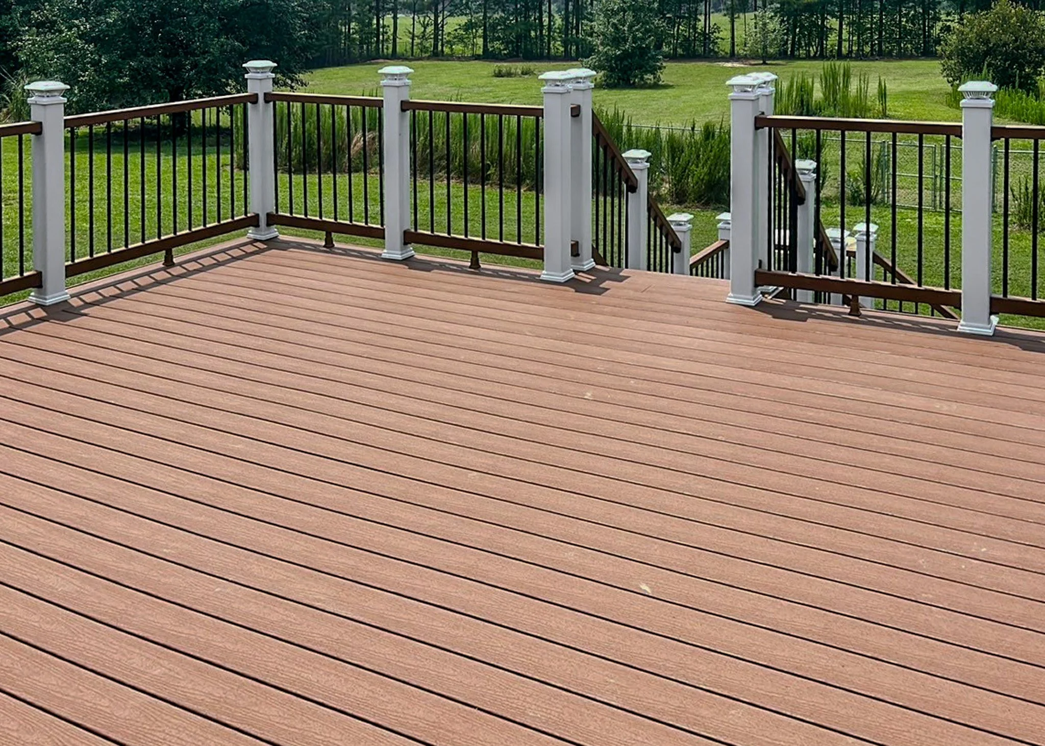 An elevated outdoor deck with wooden planks, black and white railing, and stairs leading down to a grassy yard with trees in the background.