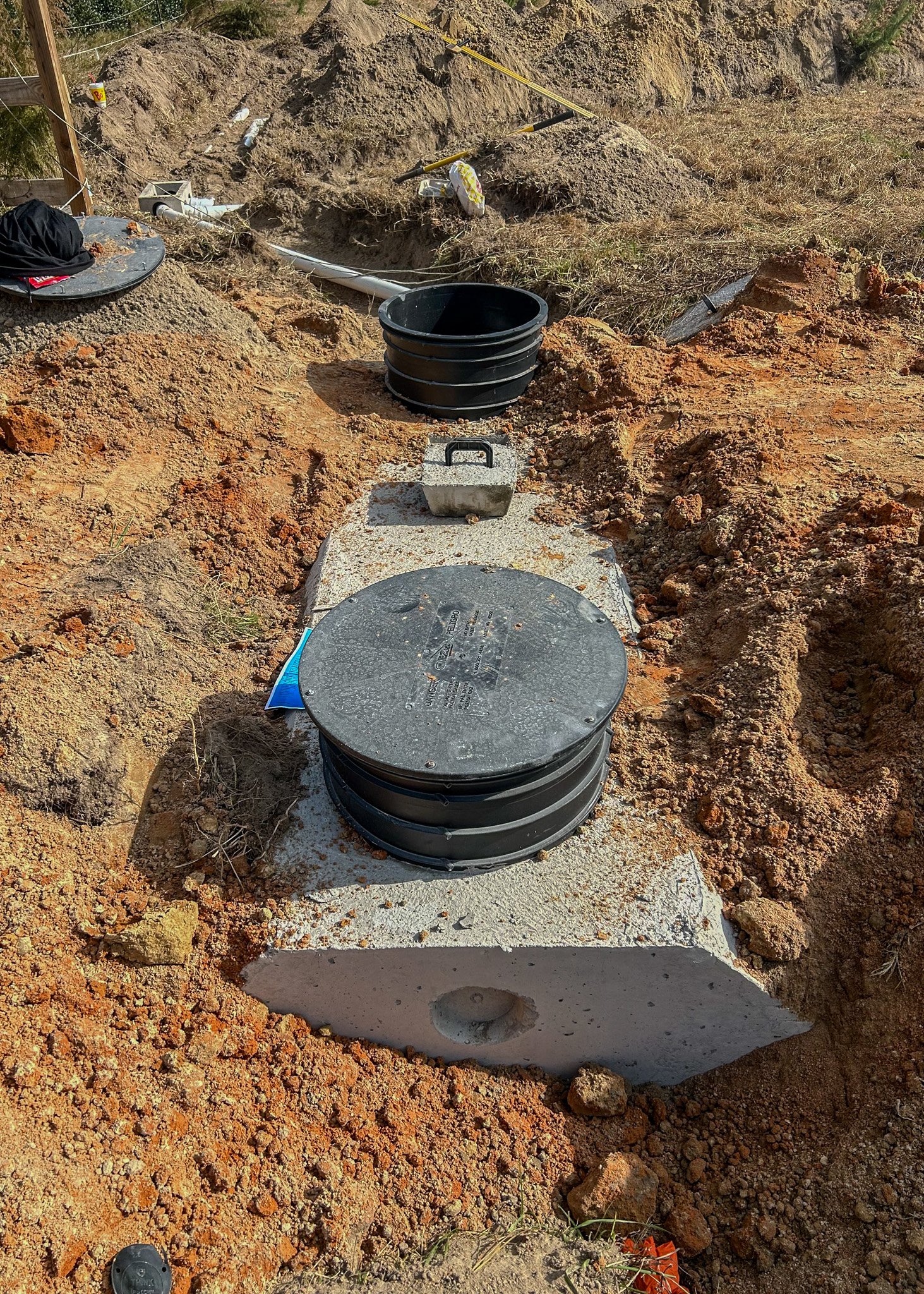 Under construction site with underground utility access covers on concrete base, black buckets, pipes, tools, and disturbed soil.