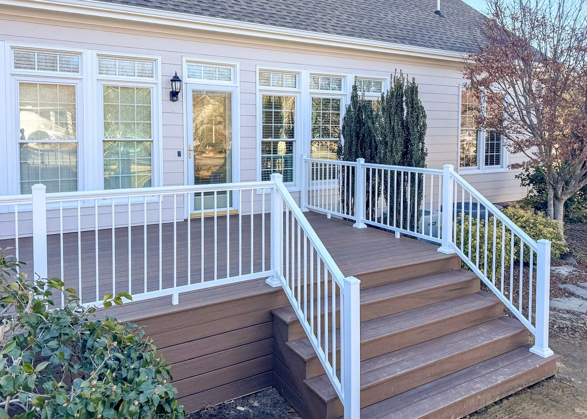 Backyard deck with white railing, wooden steps, and a sliding glass door leading into a house. There is landscaping with plants and trees, and the house has light-colored siding and multiple windows.