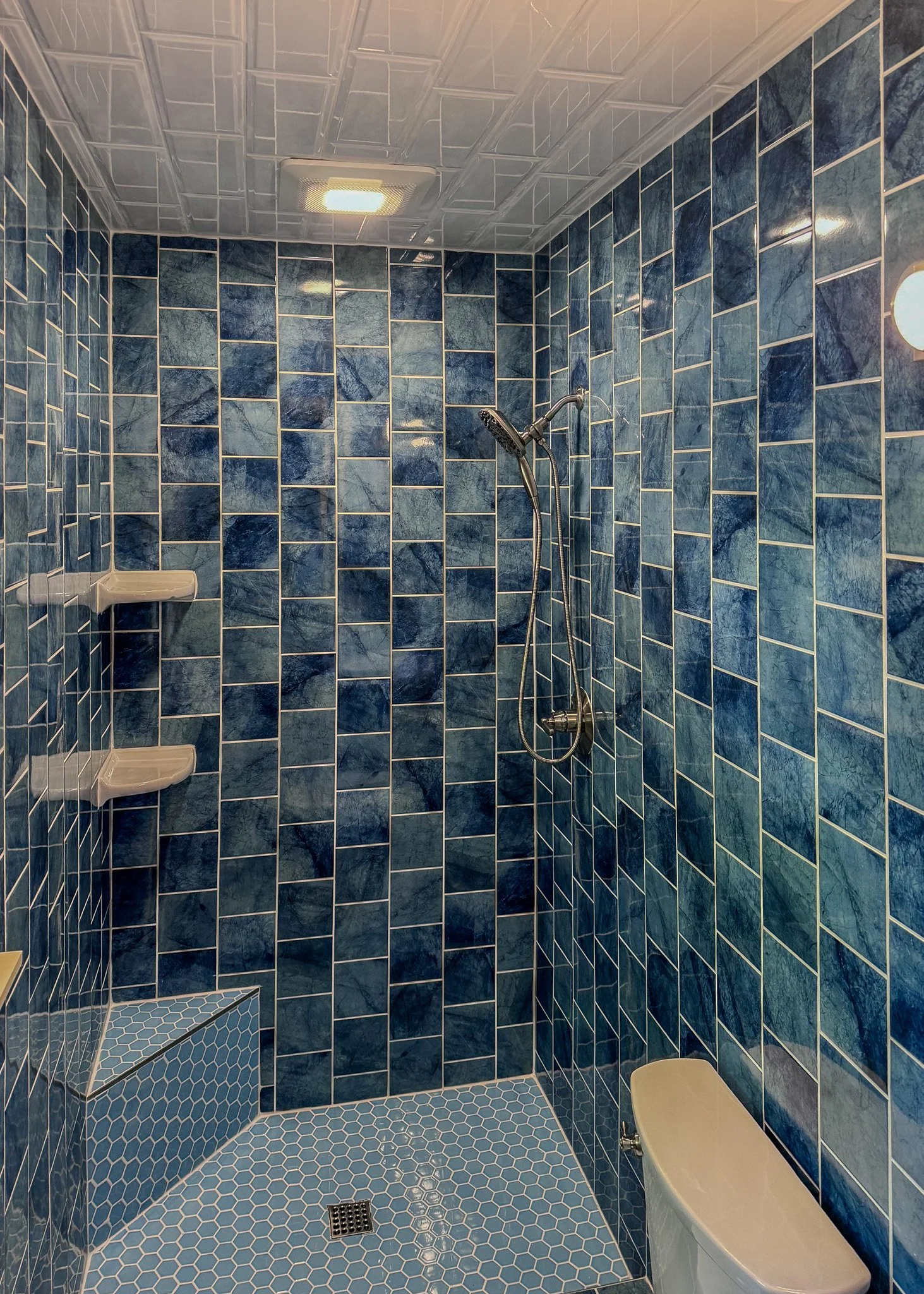 Bathroom shower area with blue tiles, white corner shelves, a handheld showerhead, a ceiling light, and a toilet.