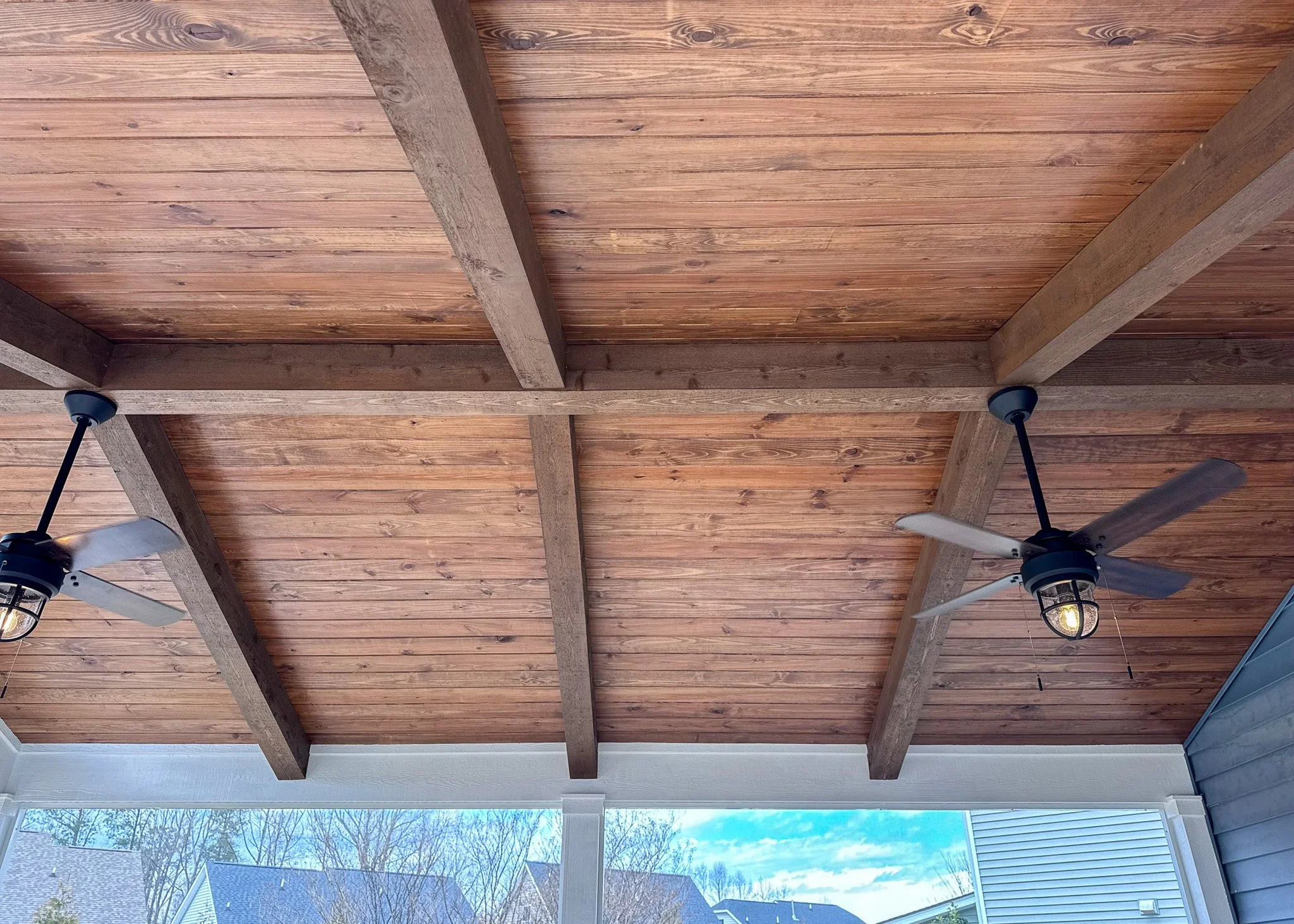 Wooden patio ceiling with exposed beams and ceiling fans, view of houses and trees outside.