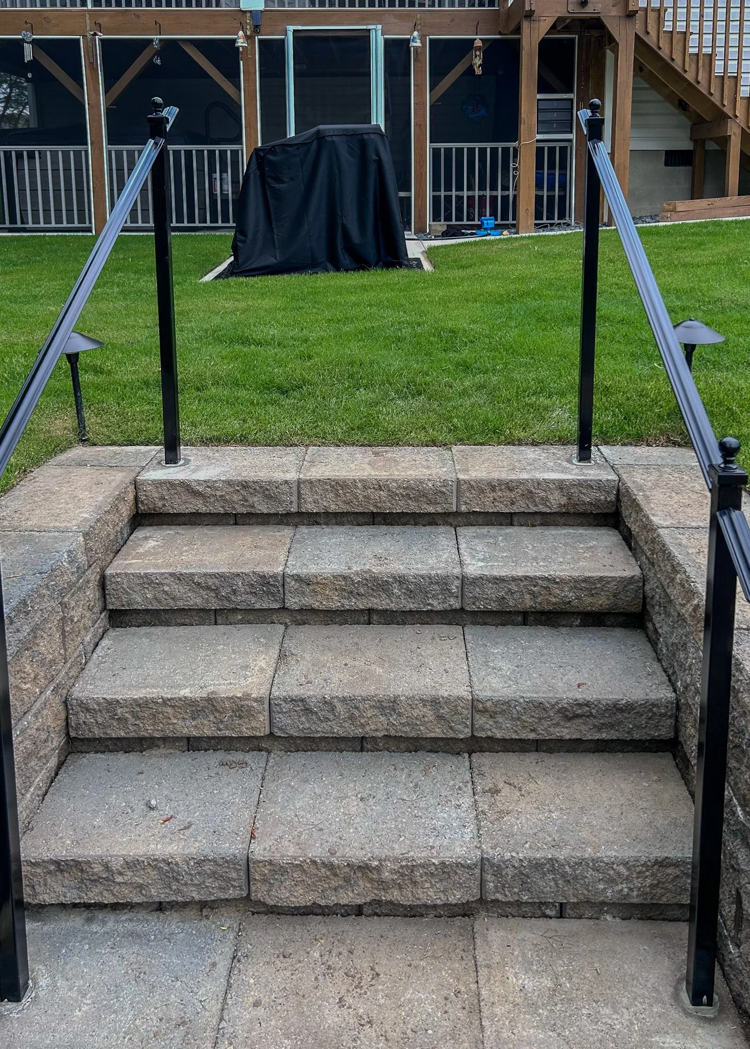 Stone steps leading up to a backyard with a grassy lawn. The steps are flanked by black metal railings. In the background, there is a deck with bean bag chairs covered with black fabric, a staircase leading to the upper level, and part of a house wit
