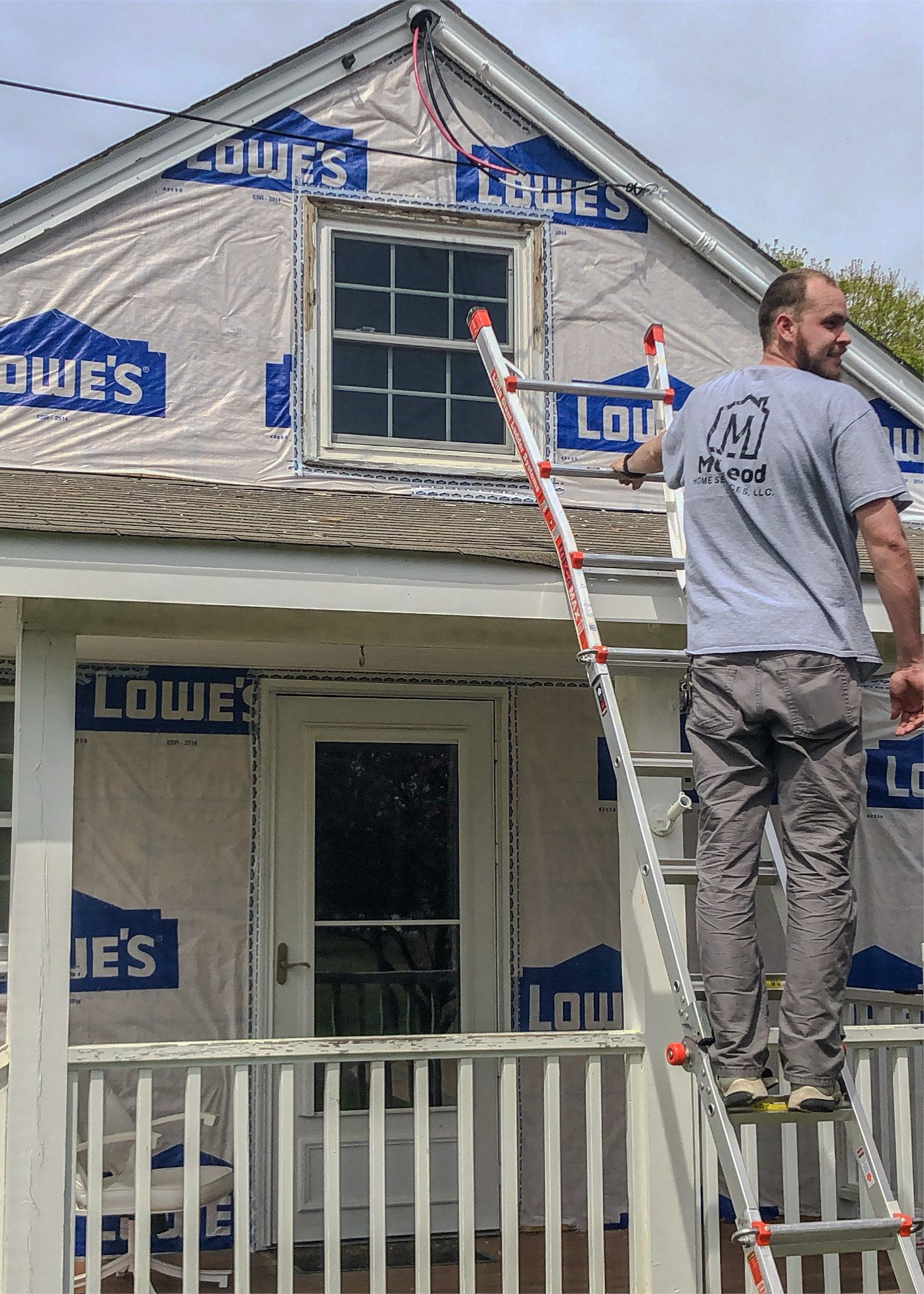 A man is working on the exterior of a house under renovation, standing on a ladder on a porch with a white railing. The house is partially covered with Lowe's branded house wrap, indicating ongoing construction or repairs.