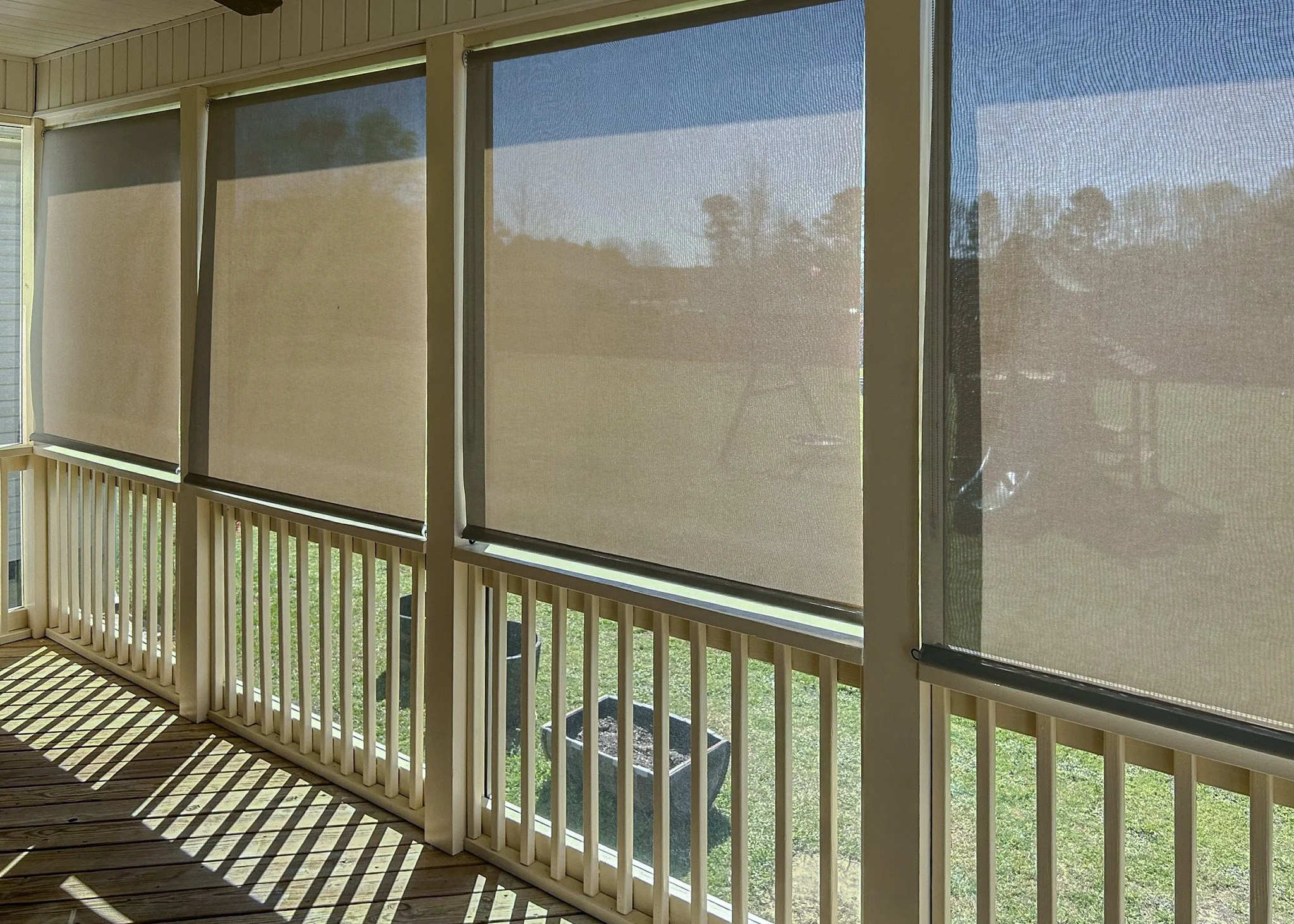 Screened porch with four window screens allowing sunlight to cast striped shadows on wooden deck flooring, showing a grassy backyard outside.