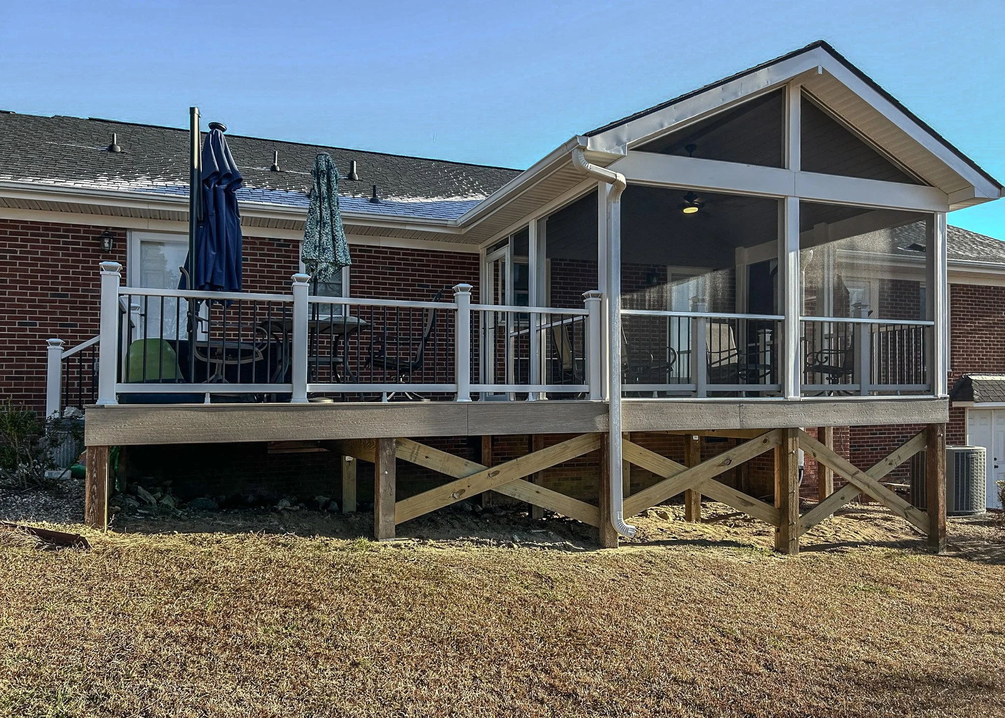 Backyard deck with patio furniture and umbrellas, attached to a brick house with a screened porch, under a clear blue sky.
