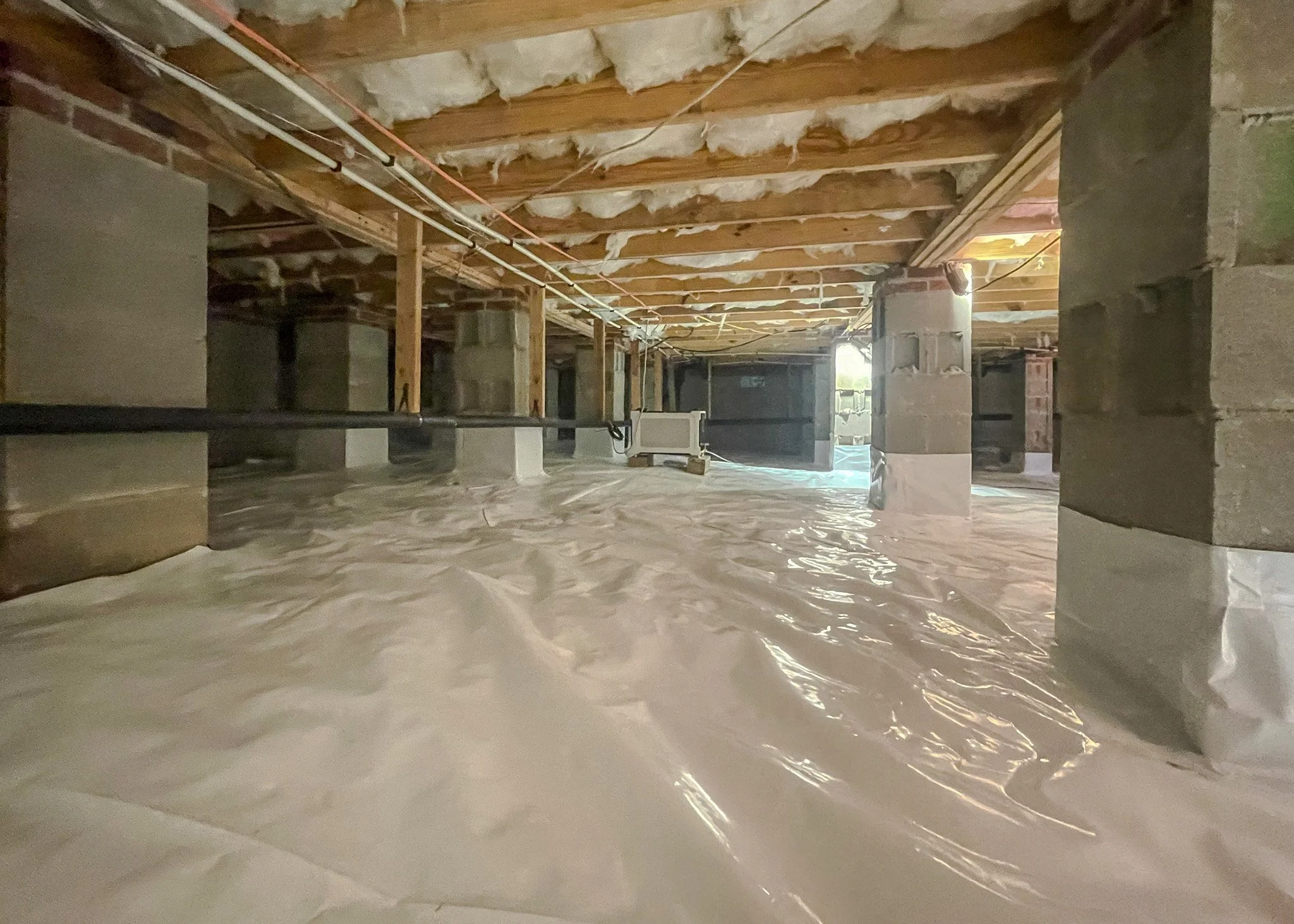 Basement with exposed ceiling joists, insulation, concrete block support columns, electrical wiring, and a plastic vapor barrier on the floor.